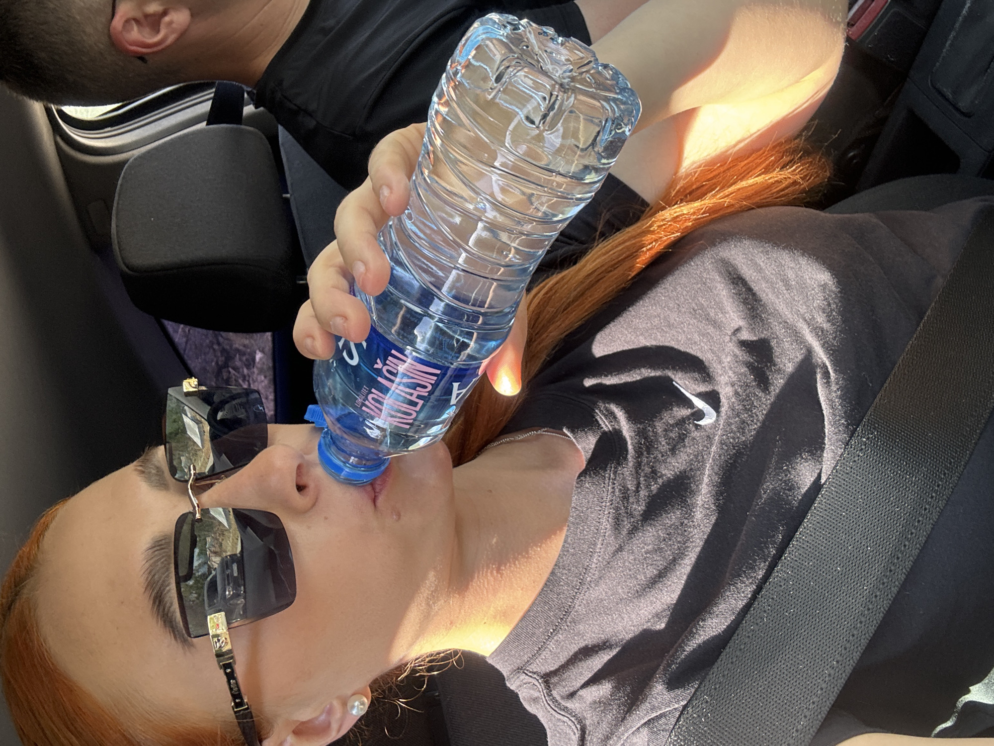 A woman is drinking from a plastic water bottle. She is wearing sunglasses and a black T-shirt with a white Nike logo, secured by a seatbelt. Another person is partially visible in the upper left corner of the frame. The setting appears to be inside a vehicle. No data indicates this image was taken near or in the city of Podlastva (Poljice), Montenegro.