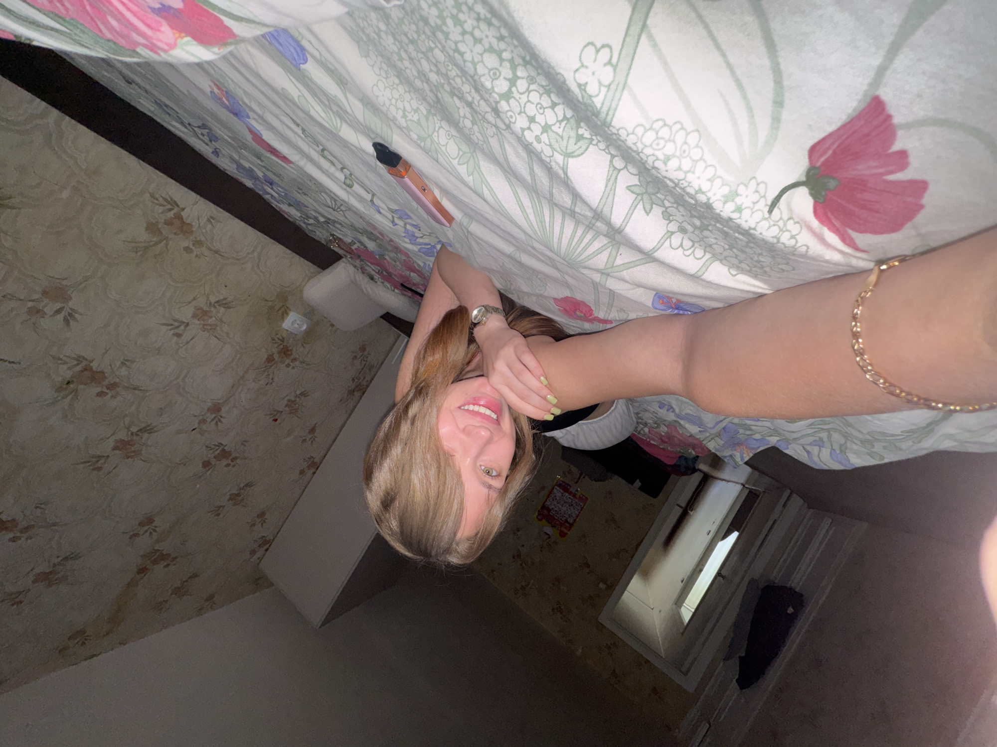 In a dimly lit bedroom, a young woman with blonde hair and light eyes is lying on a bed covered with floral-patterned bedding. She is smiling and looking at the camera, with her left arm resting on her chest. Her right arm is extended towards the viewer, with a gold bracelet on her wrist. A watch is visible on her left wrist. A pink and gold hair straightening iron lies on the bed next to her. The room's wallpaper has a floral pattern with faded colors. To the left of the bed, a doorway leads to another part of the room, where clothing is visible on the floor. The image appears to be taken in Chaikovsky, Russia.