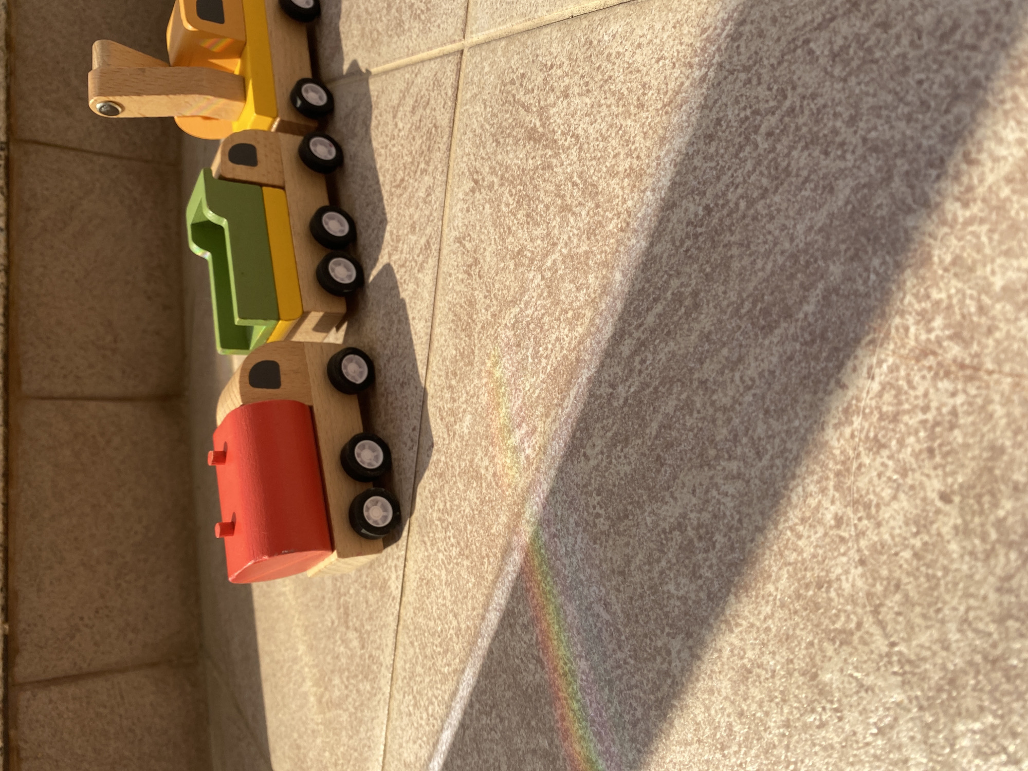 A line of three colorful wooden toy vehicles rests on a light-colored, textured tiled surface in Boreti, Montenegro. The toys are connected, forming a small train or convoy. The foremost toy is a yellow and natural wood crane or front loader, followed by a green wooden block shaped like the number "1". The third toy is a red cylindrical tank car, also made of wood. All vehicles feature small black wheels with white centers.
Strong, directional sunlight illuminates the scene, casting long, distinct shadows of the toys and other unseen objects onto the tiles, suggesting either early morning or late afternoon. The bright sun also creates a notable optical effect: a small rainbow spectrum is visible across the tiled floor, indicating that light is passing through a prism or similar refractive object before hitting the surface. The weather appears clear and sunny. No people are visible, and no specific activity beyond the arrangement of the toys is taking place. The tiled surface suggests an outdoor patio or an indoor space with natural light. The only visible text is the number "1" formed by the green toy.