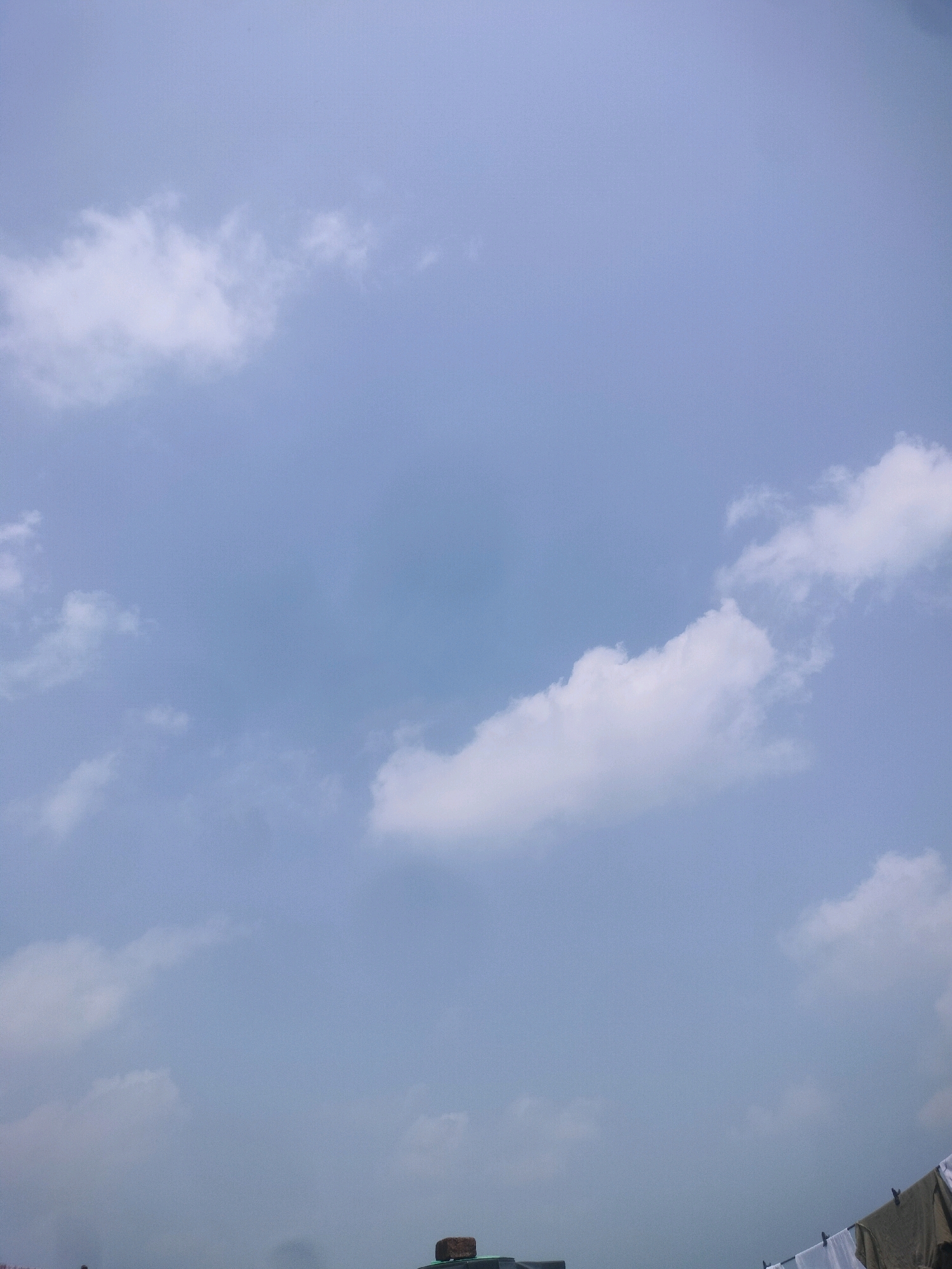 The image shows a pale blue sky with scattered white clouds. The bottom of the image reveals the rooftop of a building, with some laundry hanging on a clothesline. The scene appears to be set in Raiganj, India, given the context. There are no people visible in the image. The focus is on the sky and the rooftop details.