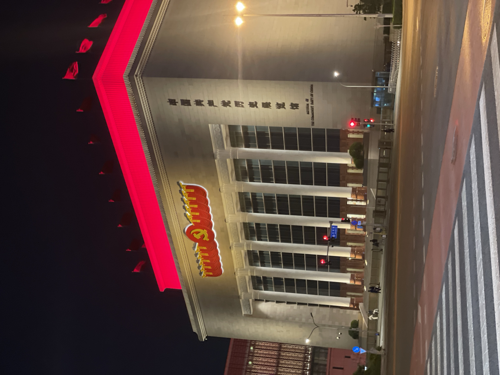 The image shows the Museum of the Communist Party of China at night. The building has a neo-classical facade and is illuminated. A red neon sign with the Communist hammer and sickle logo is visible on the side of the building. Strings of red flags run along the top of the building. The building's name is inscribed in Chinese characters and English. A street with crosswalks is in the foreground. Streetlights and traffic signals are present. The location is likely within Changping District, China, but further information on exact city and street address not available from image.
