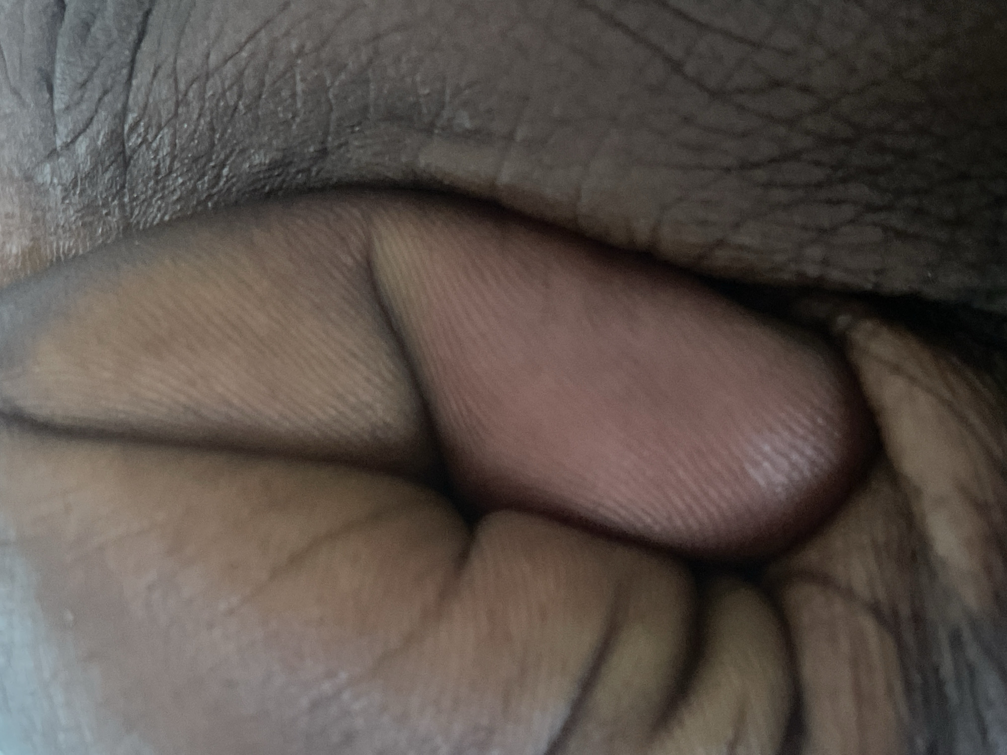 The image is a close-up, detailed view of human skin, specifically focusing on the texture and creases of fingers and knuckles. The skin appears to be of darker complexion, with visible lines and ridges characteristic of fingerprints and the natural folds of the hand. One finger is prominently featured, with its tip showing a reddish hue, possibly from light reflection or a slight blush of color. The surrounding areas reveal the texture of other fingers or parts of the hand, with shadows and highlights emphasizing the contours and depth of the skin. The image is abstract, with no discernible objects, scene, or setting beyond the human anatomy itself. The activity or event is not evident, as the focus is purely on the static texture of the skin. There are no visible cues for time of day, weather, location, or emotions. No text is present in the image. The provided location context, Rukuba Road, Nigeria, is not visually represented in the image itself.