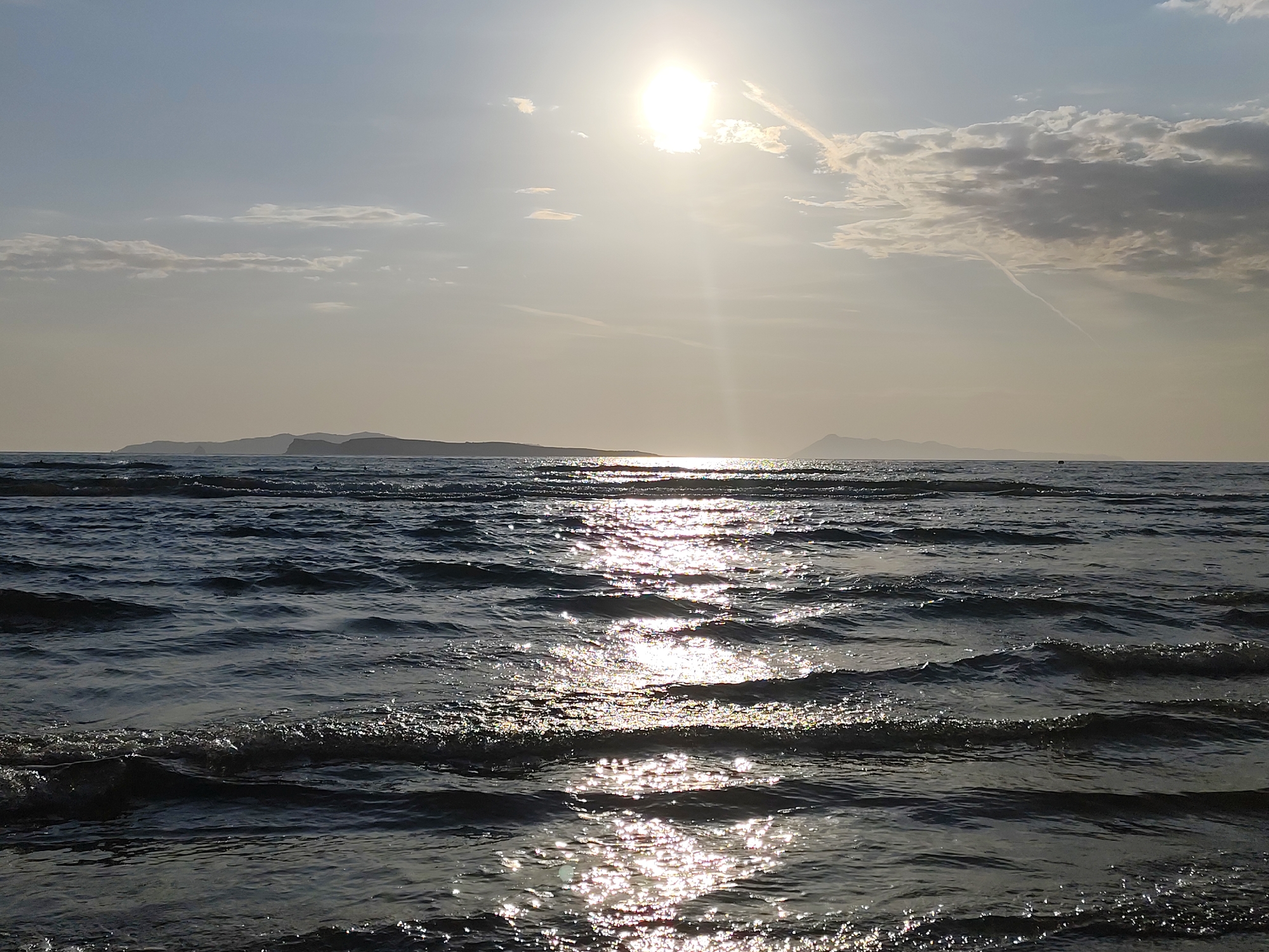 The image captures a serene scene at the beach in Agios Stefanos, Greece. The sun, high in the sky and partially obscured by thin clouds, casts a shimmering reflection on the surface of the water. The waves, gently lapping towards the shore, are illuminated by the sunlight, creating a sparkling effect. In the distance, a landmass is visible, silhouetted against the horizon. The overall color palette is subdued, with shades of blue, gray, and gold predominating, lending a tranquil atmosphere to the scene.
