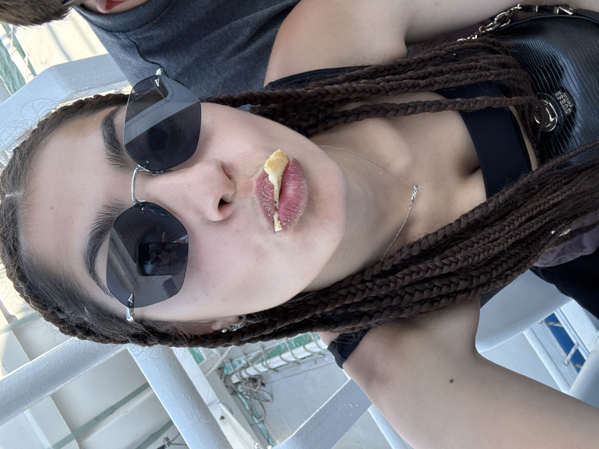 The image is a close-up selfie of a young woman on a boat. She is wearing large, dark sunglasses and has her mouth open with a piece of food on her lips. Her hair is styled in long, dark braids. She is wearing a black top and a silver necklace. A black purse with a gold chain is visible in the foreground. The background appears to be part of a boat with white railings and blue water. The location is identified as the city of Prinos, Greece.