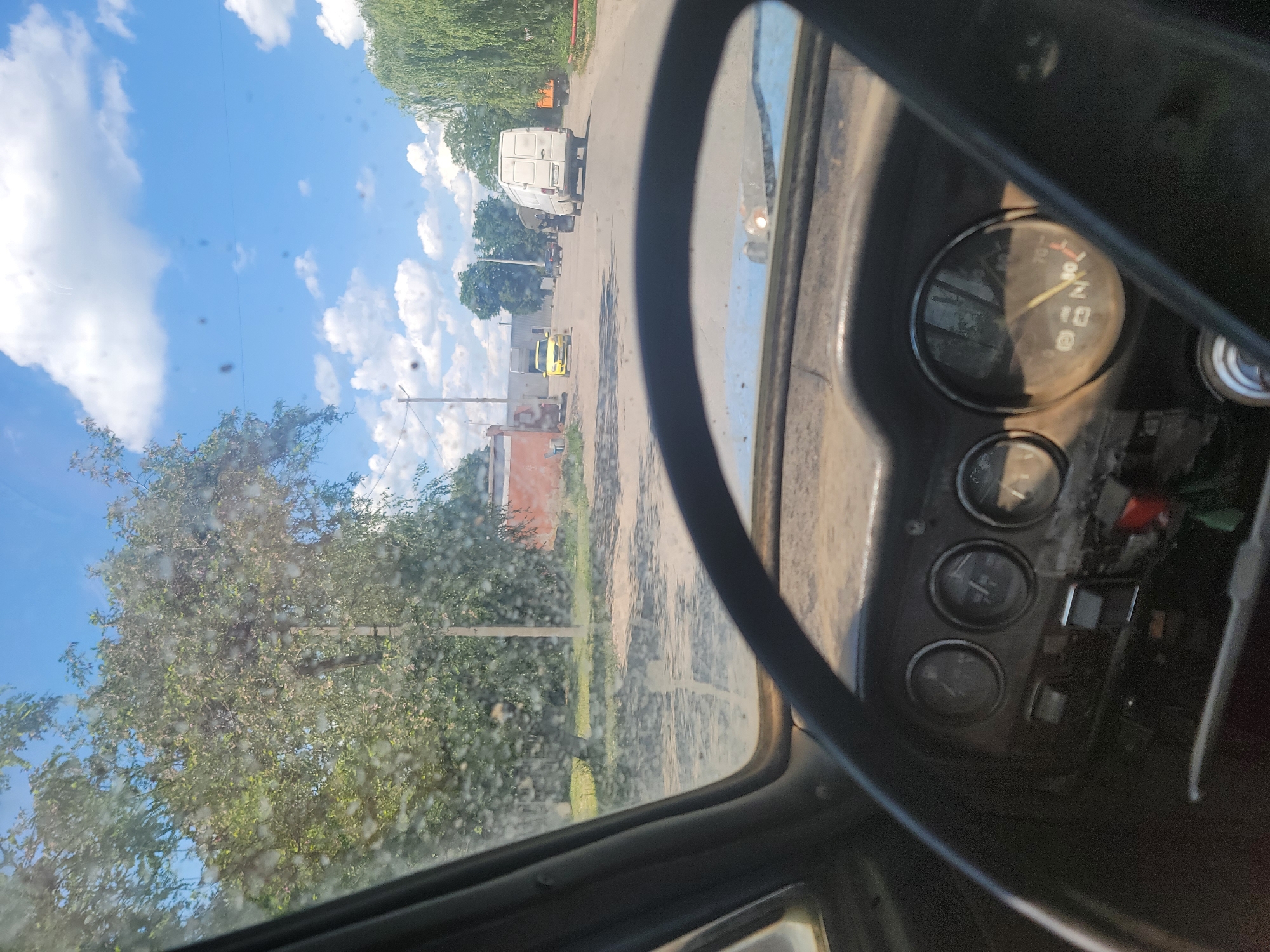 The image, taken from inside a vehicle, shows a street scene in Pereshchepyne, Ukraine. The foreground is dominated by a dirty steering wheel and dashboard with multiple gauges, indicating the vehicle is likely an older model. The windshield is visibly dirty and speckled with what appears to be water spots or dust.
The view through the windshield reveals a paved road leading into the distance. To the left of the road, there is a building with an orange facade and a tall pole. A large, leafy tree with dappled sunlight filtering through its branches stands prominently on the left side of the road, adjacent to the building. Further down the road, several vehicles are visible: a yellow car is parked on the side, followed by a white van in motion, and then a dark-colored truck. To the right of the road, a weeping willow tree with long, drooping branches shades the scene. The sky is bright blue with scattered white clouds. The overall impression is a sunny day in a rural or semi-urban setting.