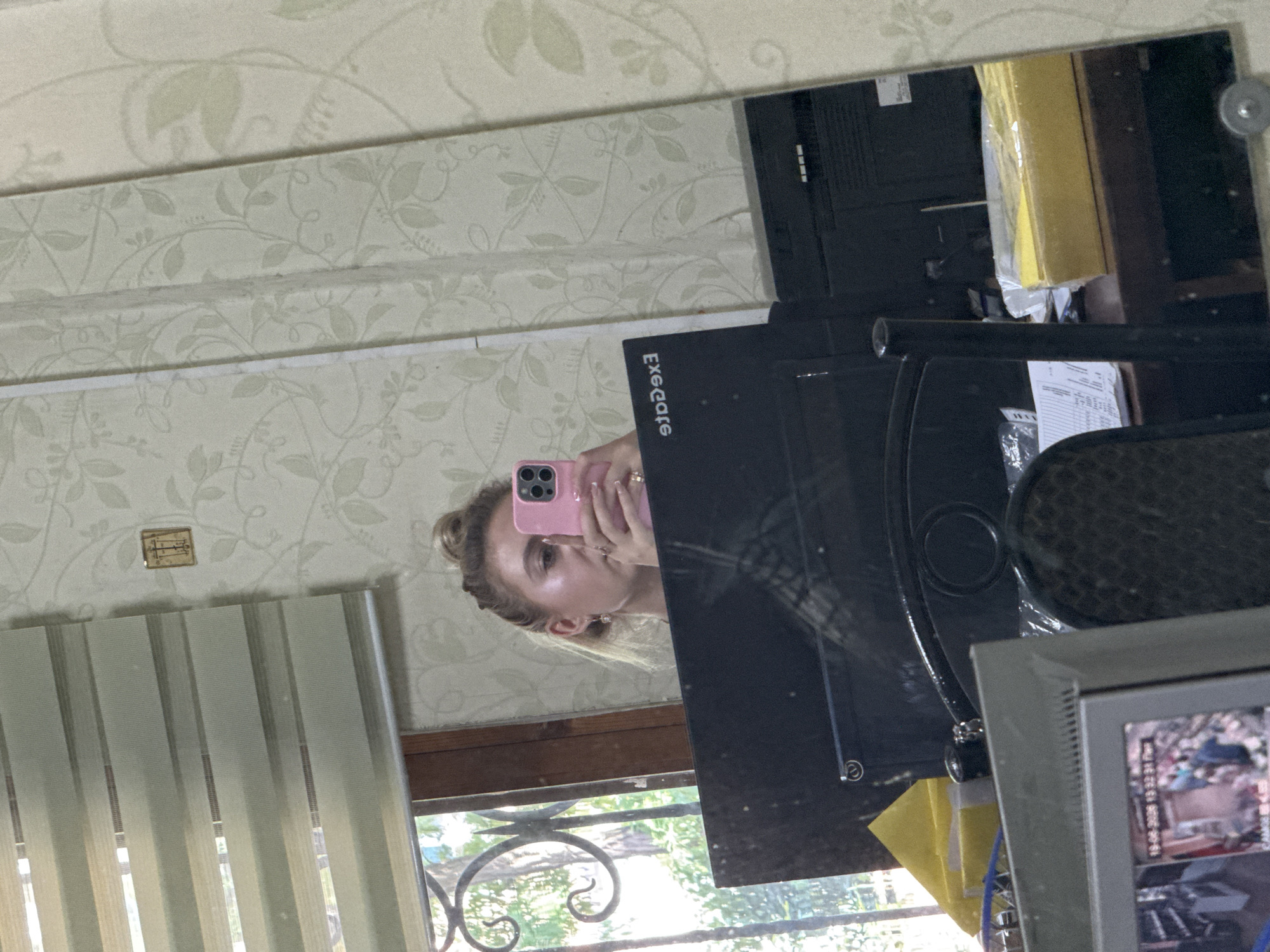A young woman with blonde hair tied in a bun is reflected in a mirror, taking a selfie with a pink smartphone. She is wearing a dark top and jewelry. The background visible in the mirror includes a window with blinds, a wall with floral wallpaper, and a desk with computer monitors and office supplies. Another reflection shows a close-up of a computer monitor displaying a security camera feed with a timestamp.