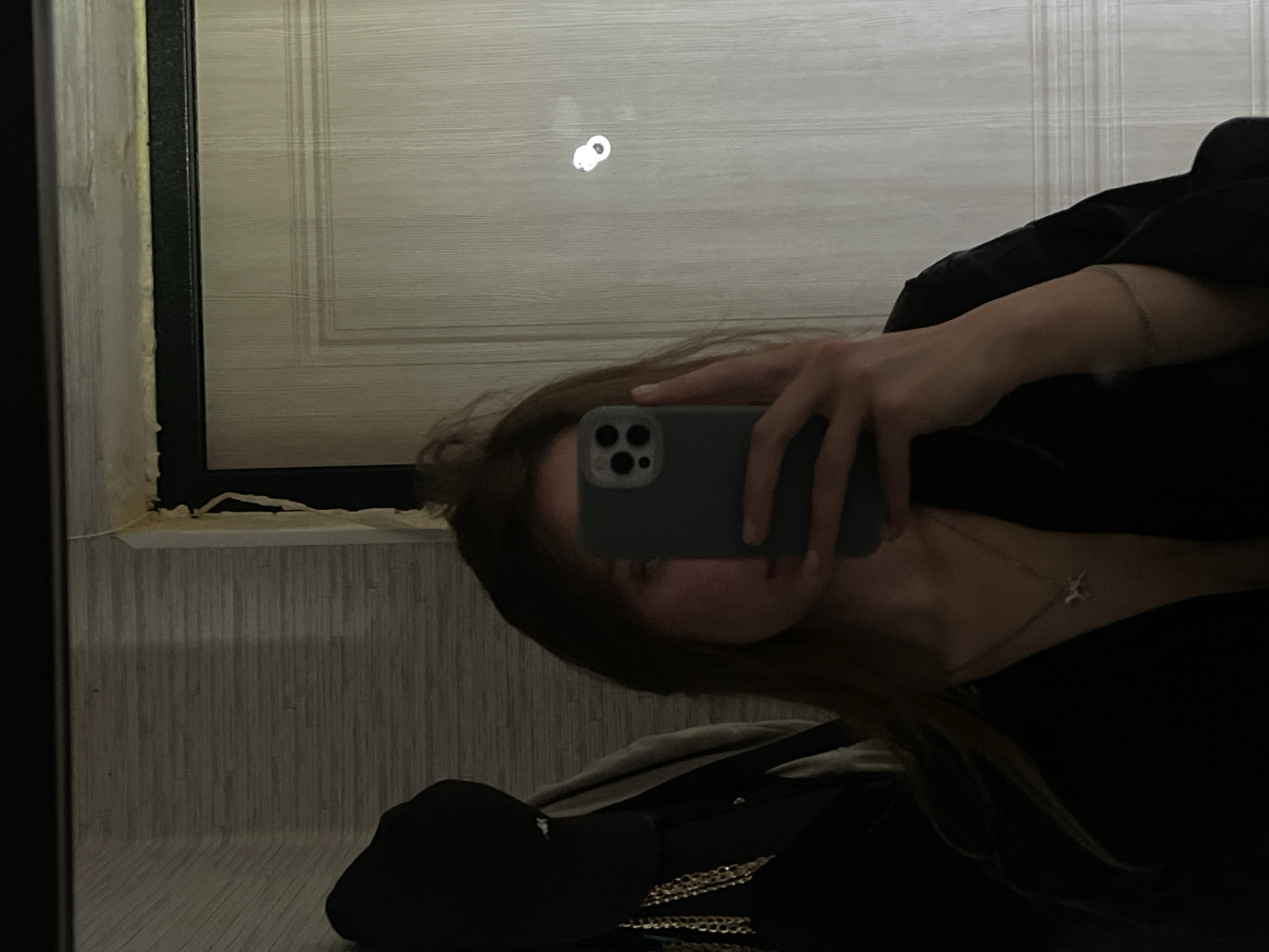 A person is taking a mirror selfie in an indoor setting, possibly a hallway or closet. The person is holding a smartphone in their right hand, obscuring their face. They are wearing a black top and a silver necklace with a small pendant. To their left, a dark object, possibly a hat or bag, hangs on a hook. The background consists of a wall with textured wallpaper and a door with white paneling. The lighting is dim, creating shadows and highlighting certain areas. There is also a visible piece of foam insulation around the doorframe, suggesting recent construction or repair work. The overall impression is a casual, personal moment captured in a domestic environment. There is no information in the image to ascertain its location in Chaikovsky, Russia.
