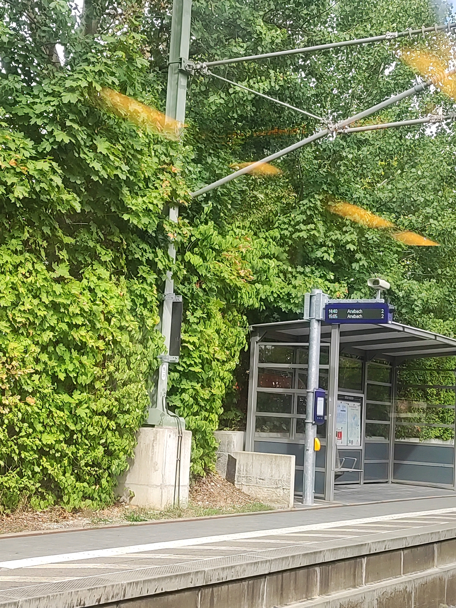 The image shows a bus stop shelter with a digital display sign in Roßtal, Germany. The display shows departure times and destinations, with "14:40 Ansbach 2" and "15:05 Ansbach 2" visible. The bus stop is situated next to a railway track, indicated by the overhead electrical lines and the platform in the foreground. Lush green trees and ivy cover the background, creating a natural, slightly overgrown setting. A concrete post supports the display sign and overhead wiring. The shelter itself has glass panels and a metal frame. The lighting suggests it is daytime, with some sunlight filtering through the trees, creating yellow reflections on the overhead wires.