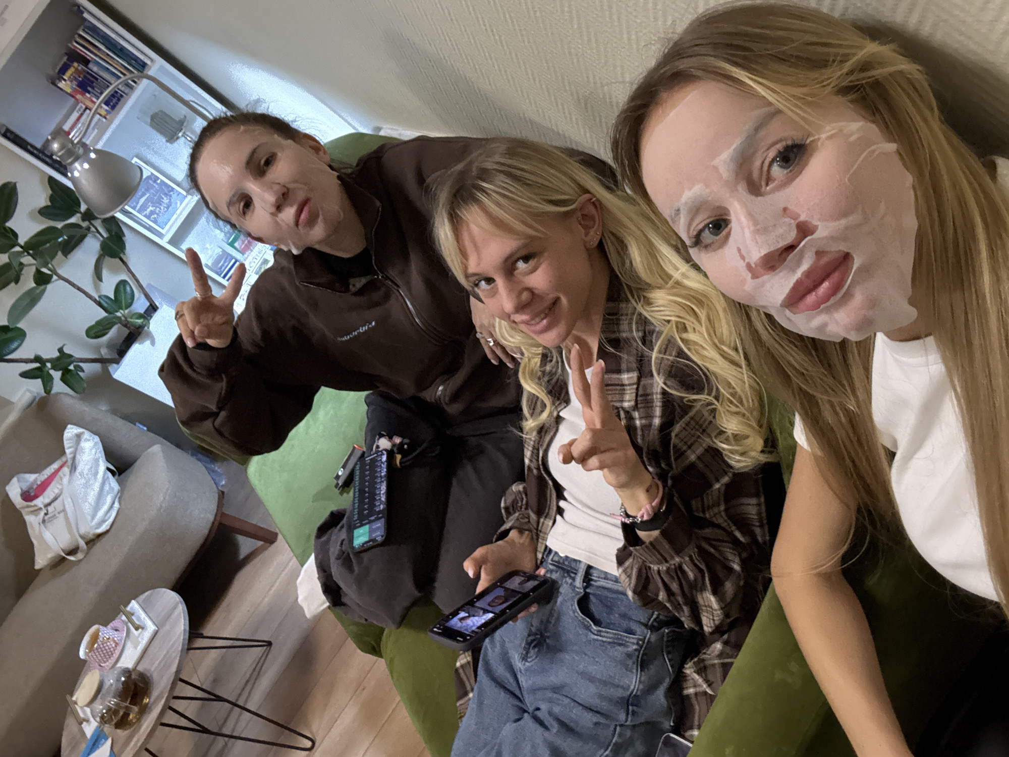 Three young women are posing for a selfie in an interior setting, likely an apartment in Moscow, Russia. The woman on the left has short brown hair and is wearing a brown sweatshirt. She is making a peace sign and has a facial mask on. The woman in the center has blonde hair, is wearing a plaid shirt over a white top, and is holding a phone. She is also making a peace sign. The woman on the right has long blonde hair and is wearing a white top. She has a facial mask on. They appear to be sitting on a green couch. A coffee table with teacups and a teapot is visible to the left, along with a gray sofa and a bookshelf. Two phones and a small remote control are visible on the couch to the left. The overall scene suggests a casual, domestic environment, possibly a girls' night in.