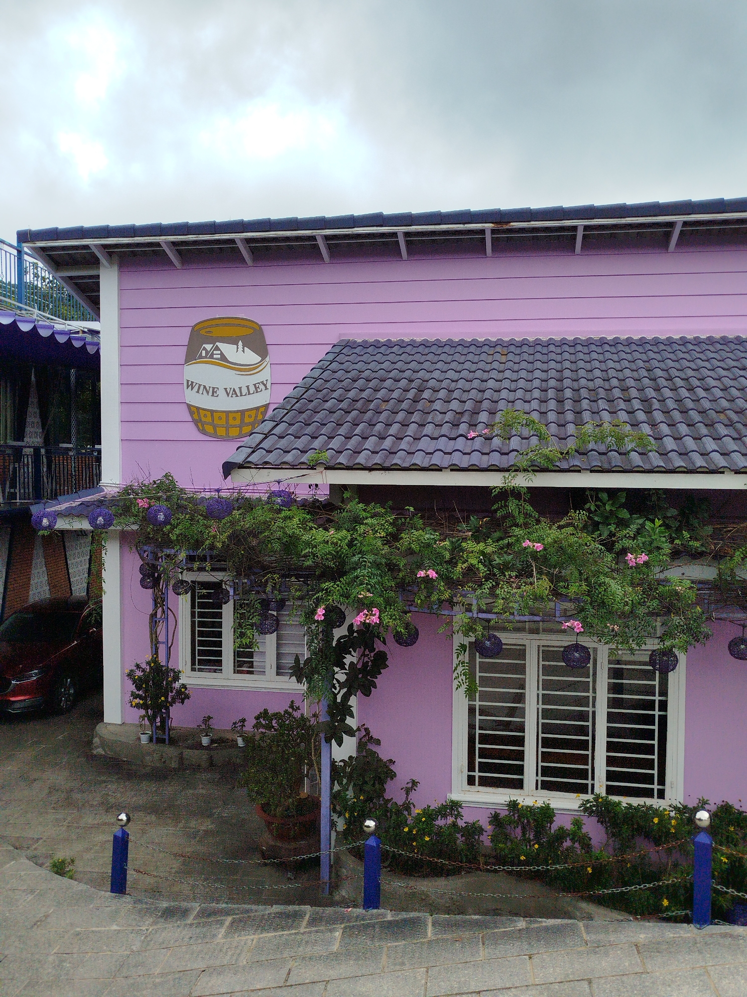 A pastel pink building, branded "Wine Valley," stands under a grey, overcast sky. The building features a dark tiled roof, white-framed windows with horizontal bars, and is partially covered in lush green vines with small pink flowers. A circular sign depicting a barrel and mountains is prominently displayed on the upper portion of the building's exterior. In the foreground, a paved walkway curves around blue posts connected by a chain, bordering a garden with various plants and flowers. A red car is visible parked to the left of the building. The scene is likely located in Da Lat, Vietnam, given the architectural style and common use of pastel colors in local establishments.