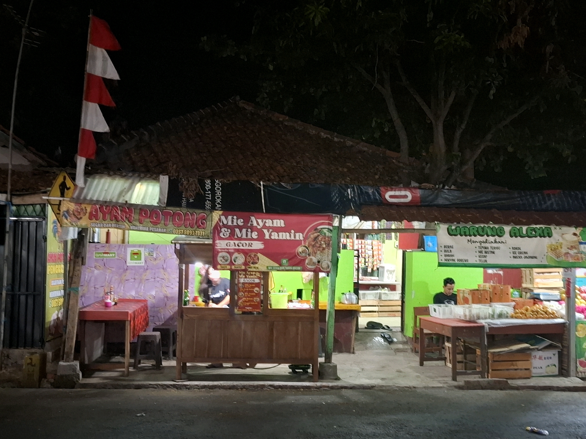 The image depicts a street food stall in Bojong, Indonesia, at night. The stall is illuminated by artificial lighting, with a prominent red and white Indonesian flag displayed vertically on the left. The stall appears to be selling noodles and chicken dishes, with signage that reads "MIE AYAM & MIE YAMIN" and "AYAM POTONG". To the right, another section of the stall, labeled "WARUNG ALEXA", sells various goods including what appear to be fruits and packaged items. A person is seated at a table in front of the "WARUNG ALEXA" section, looking down at something on the table. Another person, a woman, is visible behind the counter of the noodle stall, engaged in activity. The surrounding environment suggests a local, informal commercial area.
