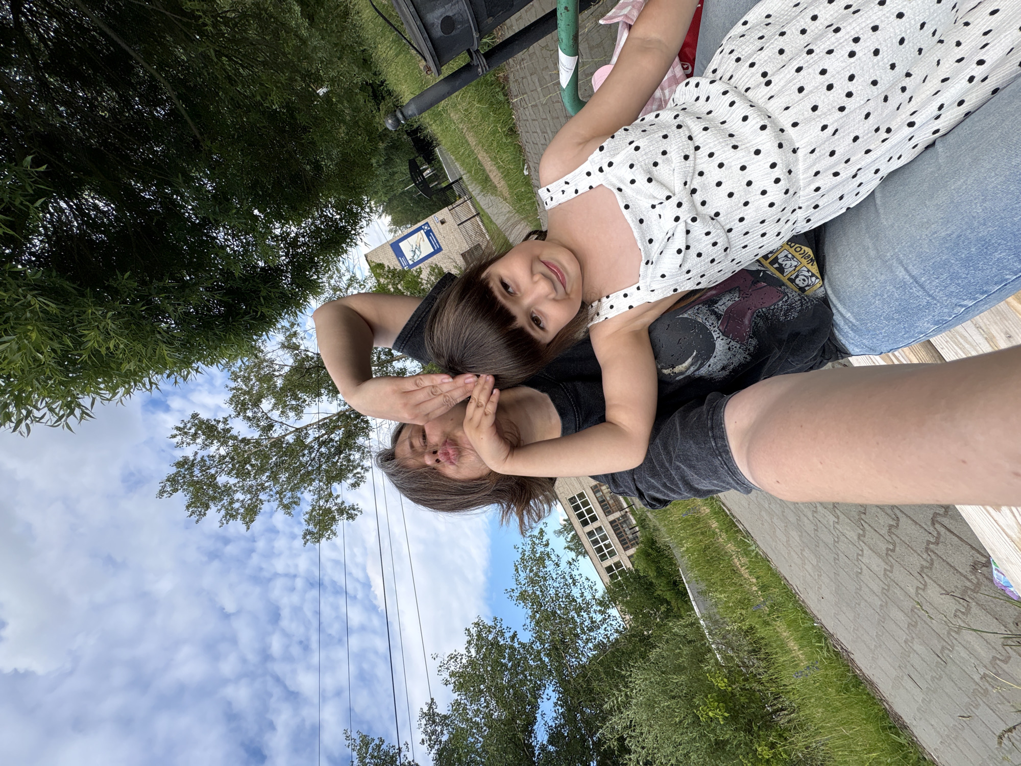 In Chaikovsky, Russia, two individuals, an adult and a child, are captured in a selfie on a paved pathway beside a grassy area. The adult, with their arm raised and hand near their face, appears to be making a kissing gesture. They are wearing a dark t-shirt. The child, wearing a white and black polka dot dress, is seated next to the adult, smiling towards the camera. Behind them, a building with a visible sign is situated amongst trees. The sky above is partly cloudy.