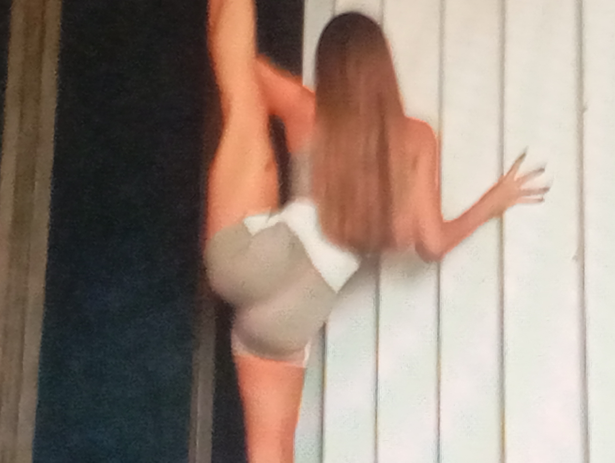 The image shows a woman with long, brown hair in a split, leaning against a white, vertical paneled wall. Her body is positioned with her left leg extended upwards and her right leg bent. She is wearing a light-colored, form-fitting garment with a white accent. Her right arm is extended towards the wall, with her fingers visible. The background includes a dark, vertical element on the left side of the image. The scene appears to be indoors or in a covered outdoor space, possibly a studio or a stage, given the lighting and the backdrop. The specific location in Sofiivska Borshchahivka, Ukraine, cannot be determined from the visual information alone.