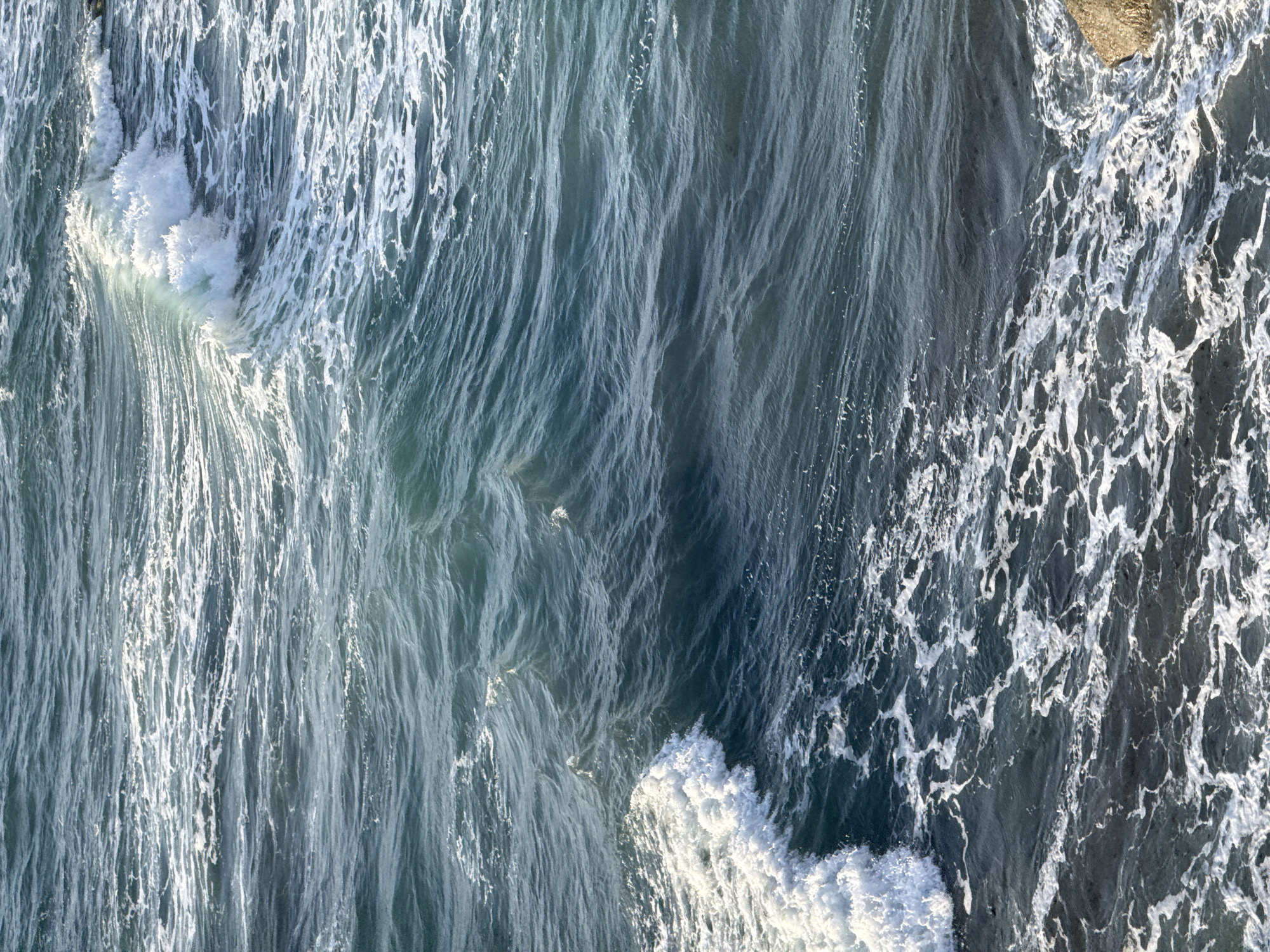 The image depicts a close-up, high-angle view of the ocean off the coast of Sopela, Spain. The water is a deep, dark blue-green, with light reflecting off the choppy surface. White foam crests are visible as waves break and recede, particularly in the lower left and upper right portions of the image. A large, light brown rock is partially submerged in the lower right corner, near the shore. The scene is natural and devoid of any human presence or man-made structures, focusing solely on the dynamic movement and texture of the sea.