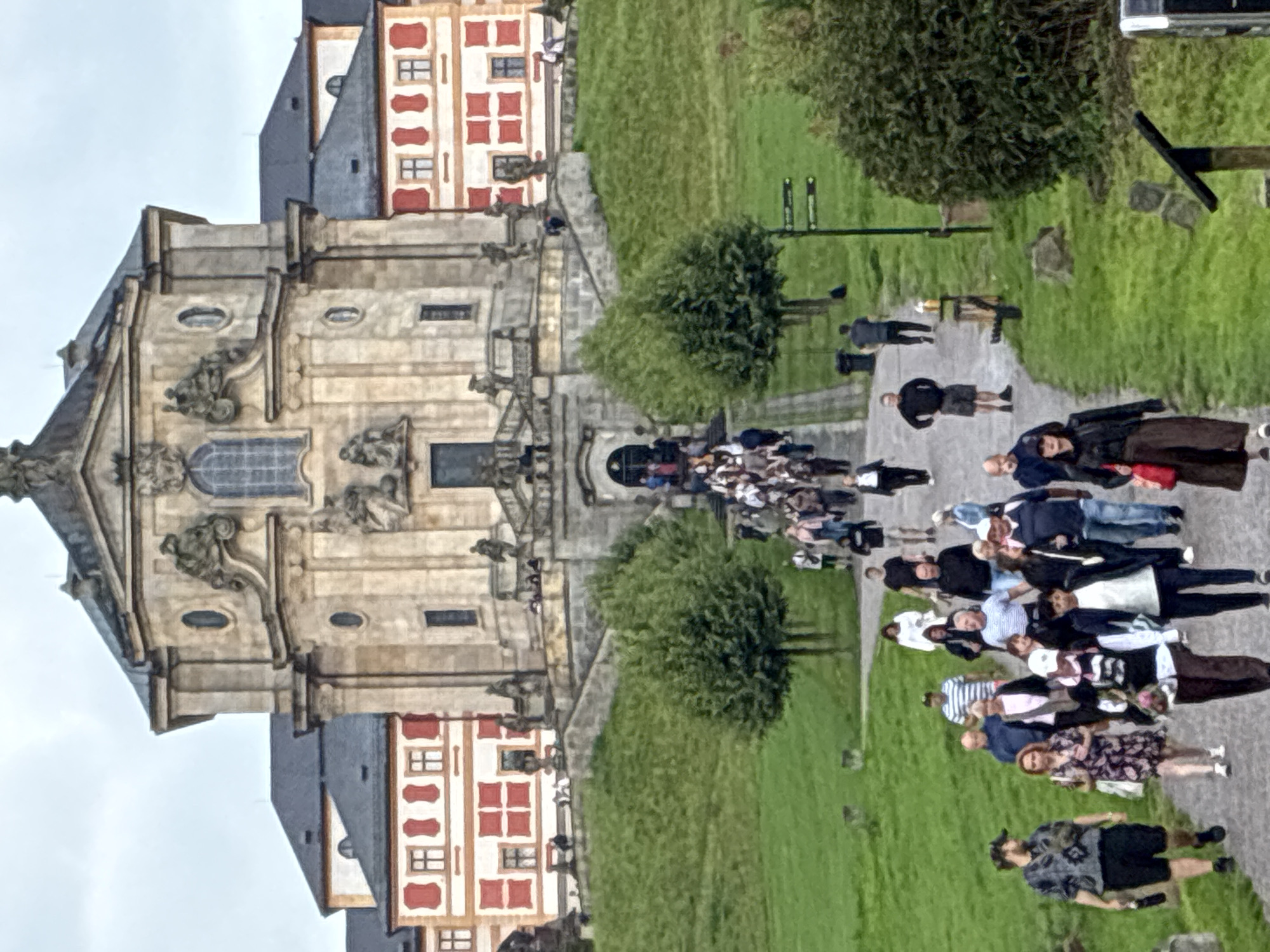 The image shows the Baroque complex of Kuks Hospital in Kuks, Czechia. A crowd of tourists is walking up a path towards the main building. The building is large and ornate, with a prominent facade and red and white accents. The path is lined with green grass and trees, and a signpost is visible on the right side of the frame. The sky is overcast. The tourists are dressed in casual clothing and are of various ages and genders. Some are walking in groups, while others are walking alone. The overall scene depicts a popular tourist destination on a day with moderate weather.
