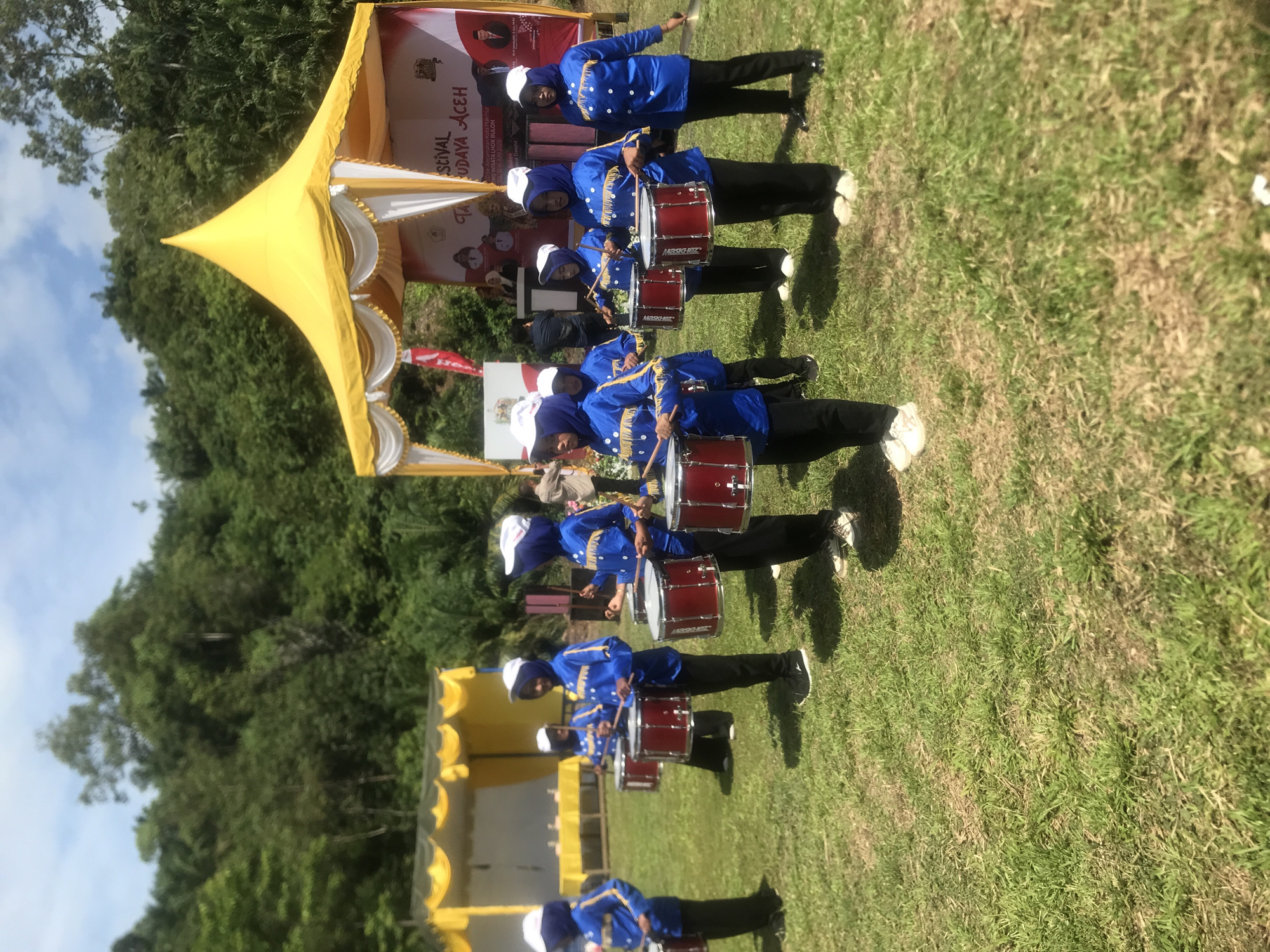 In Langkuta, Indonesia, on a sunny day, a group of young people in matching blue uniforms with white hats are performing a drumline. They are lined up on a grassy field, each holding a red drum and drumsticks. Behind them, a yellow tent is set up, with a banner that reads "FESTIVAL BUDAYA ACEH". Trees and hills form the backdrop of the scene.