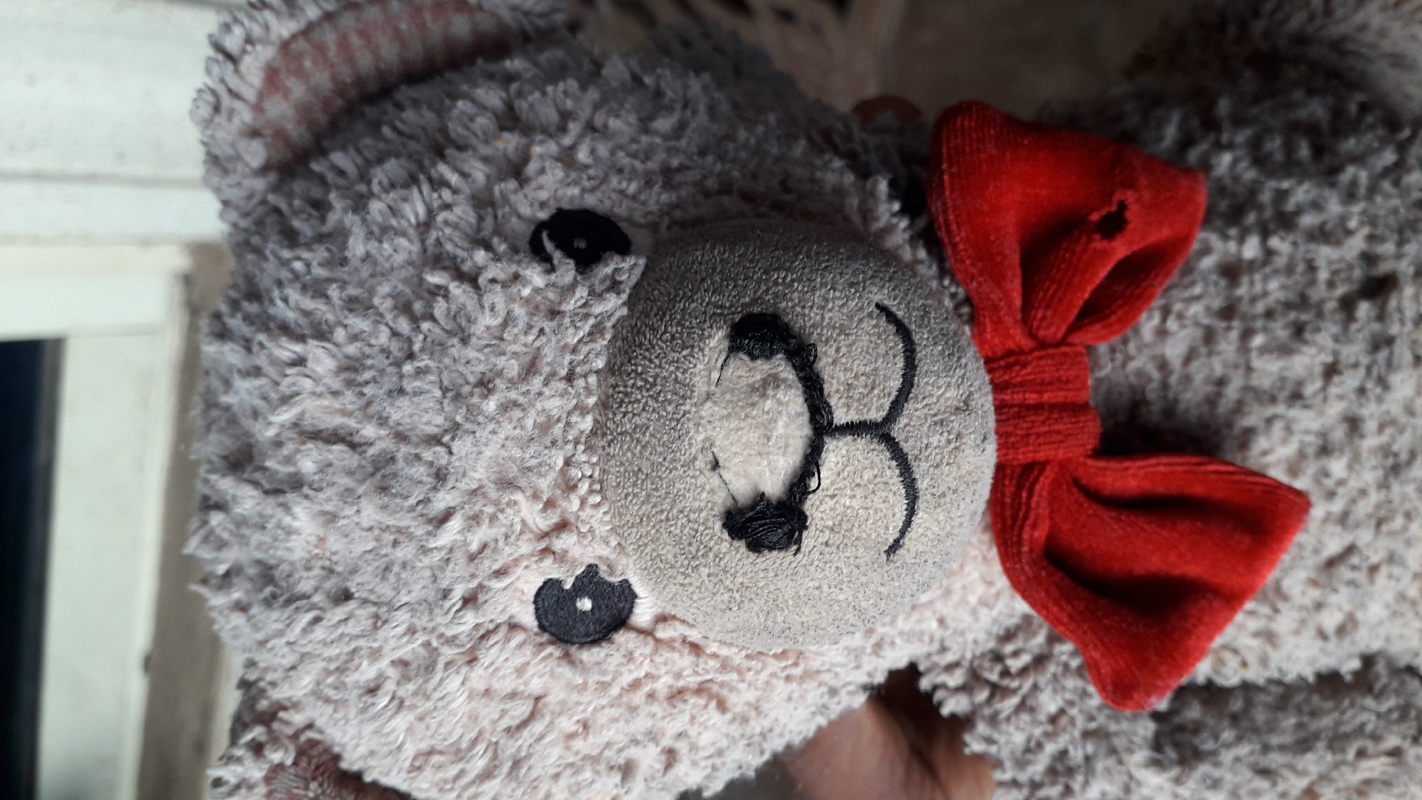A close-up image shows a worn, fuzzy, light brown teddy bear. The bear has a light tan muzzle with a stitched black nose and mouth. One of the muzzle stitches is partially undone. The bear has small, black, button eyes. A red velvet bow is attached near the bear's right ear. The bow has a small hole in the fabric. The environment is blurry and nondescript. It is not possible to confirm a location in Nkpor, Nigeria, from the visual information.