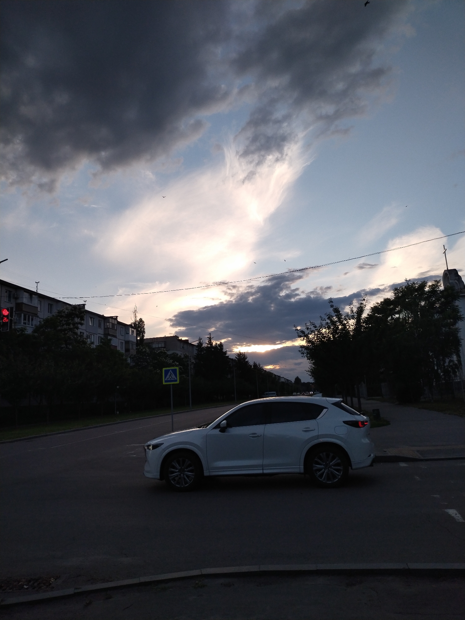 A white SUV is driving on a street in Horishni Plavni, Ukraine, during what appears to be dusk. The sky is dramatic with dark, heavy clouds mixed with lighter, wispy clouds, and a bright patch of light suggests the sun is low on the horizon. In the background, there are several apartment buildings with balconies, trees, and a building with a cross on top. A traffic light is visible on the left, showing a red signal. A pedestrian crossing sign is present near the road. Several birds are visible in the sky. The overall scene is of a city street with urban infrastructure and natural elements under a dynamic sky.