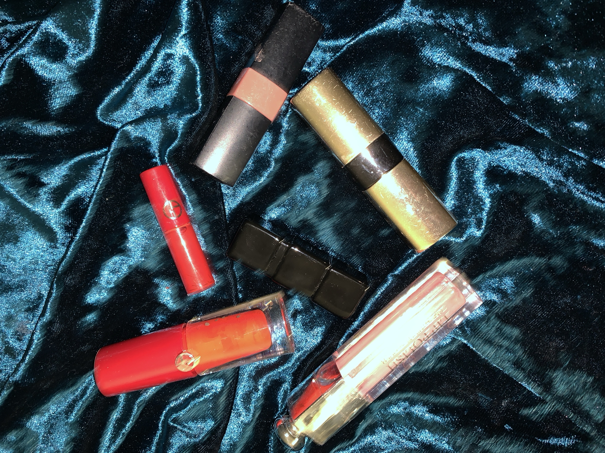 The image displays an overhead view of six cosmetic items arranged on a dark teal or petrol blue crushed velvet fabric surface. The fabric exhibits significant texture and light reflections, characteristic of velvet.
The objects, from top left to bottom right, are:
1. A clear, rectangular bottle with a bright red opaque cap, containing an orange-red liquid, consistent with nail polish or liquid lipstick. A small circular, slightly reflective emblem is affixed to the cap.
2. A small, cylindrical red tube, approximately one-third the length of the larger clear bottle, with a visible logo that appears to be a stylized 'GA' (possibly Giorgio Armani) on its side. This object resembles a miniature lipstick or lip balm.
3. A closed, rectangular black lipstick case, positioned centrally among the items.
4. A clear, elongated rectangular bottle with a silver-colored metallic cap, containing a translucent light pink or peach liquid. Text is partially visible along its side, appearing to include "LIP POLISH".
5. A black cylindrical tube with a mid-tone peach or nude colored band around its circumference. The black surface shows minor scuffs or wear. This object is consistent with a lipstick or similar product.
6. A metallic gold cylindrical tube with a black band around its circumference, similar in form to the black and peach tube. The gold surface exhibits minor wear and scuffing. This object is also consistent with a lipstick or similar product.
No individuals are present in the image. The objects are static, without any visible actions or interactions. The setting is indoors, with the exact location in the city of Unknown, Unknown, being undetermined beyond the immediate fabric surface. The lighting suggests an artificial source, creating distinct highlights and shadows on the reflective fabric and metallic surfaces of the objects.