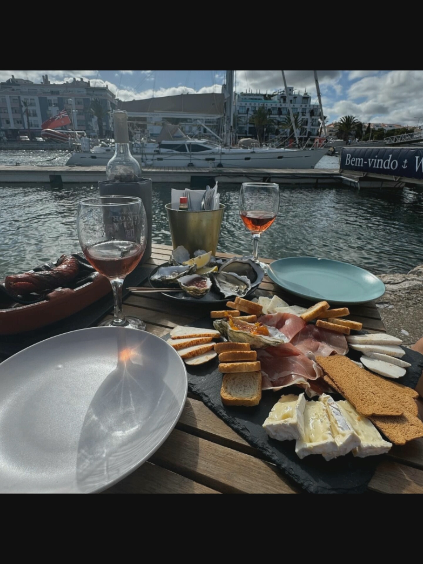 The image shows an outdoor dining scene on a wooden table overlooking a harbor. A bottle of wine, two glasses of rosé, a platter of oysters, charcuterie, cheese, and bread are arranged on the table. In the background, several yachts and buildings are visible. The location is a waterfront area in Bucharest, Romania. The weather appears to be sunny.
