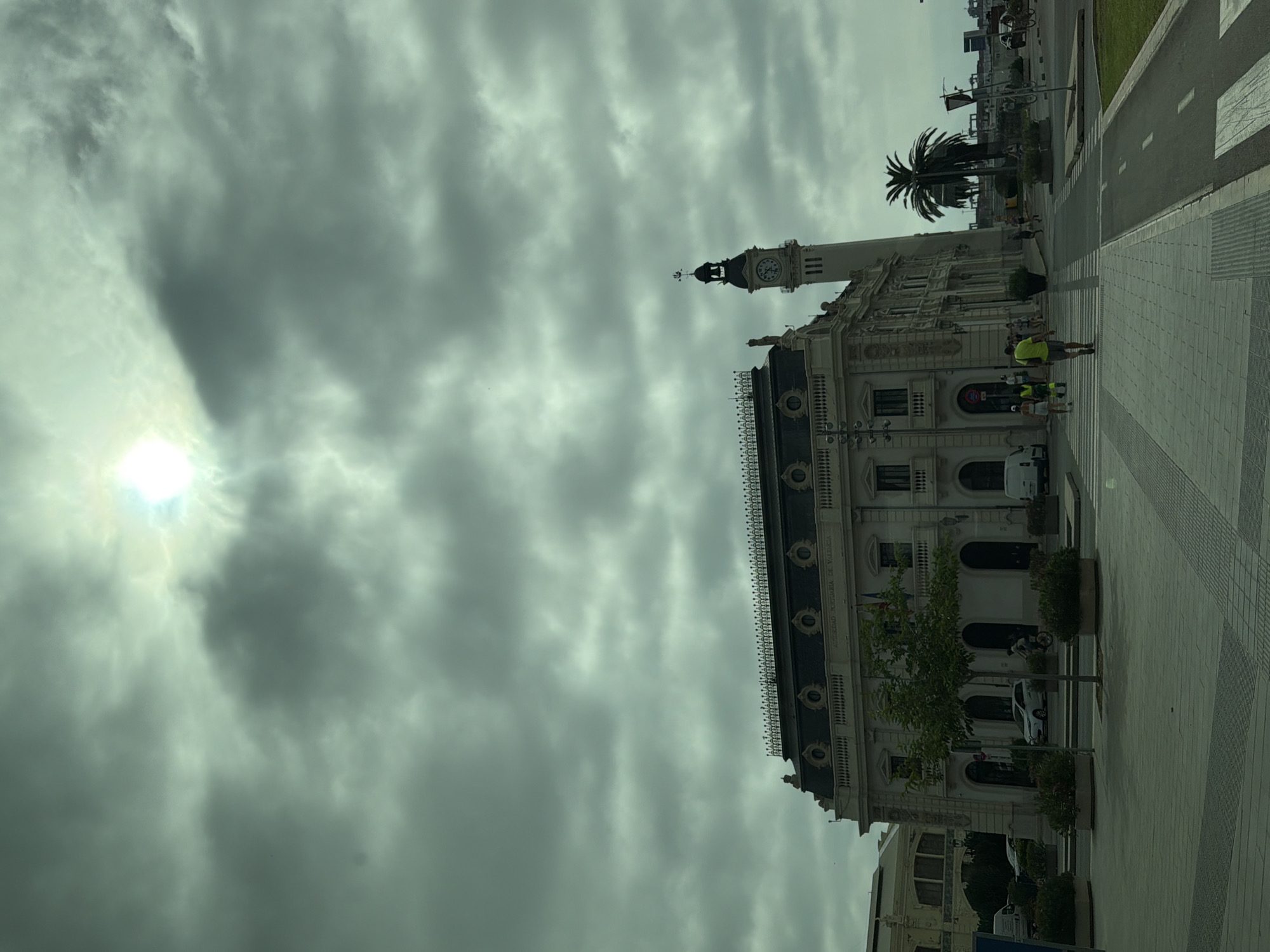 The image shows a street scene in Valencia, Spain, under a cloudy sky with the sun visible through the clouds. In the foreground, there is a wide paved walkway with planters containing bushes and small trees. A crosswalk is visible further down the walkway.
Dominating the middle ground is a large, ornate building with a dark, mansard roof and a tall clock tower. The building has classical architectural elements, including columns and arched windows. The words "AUTORIDAD PORTUARIA DE VALENCIA" are visible above the main entrance. There are several people walking on the sidewalk in front of the building and crossing the street. One person is riding a bicycle.
In the background, palm trees are visible, as well as other buildings and port infrastructure. The overall lighting suggests it is daytime, but the heavy cloud cover creates a somewhat muted atmosphere.