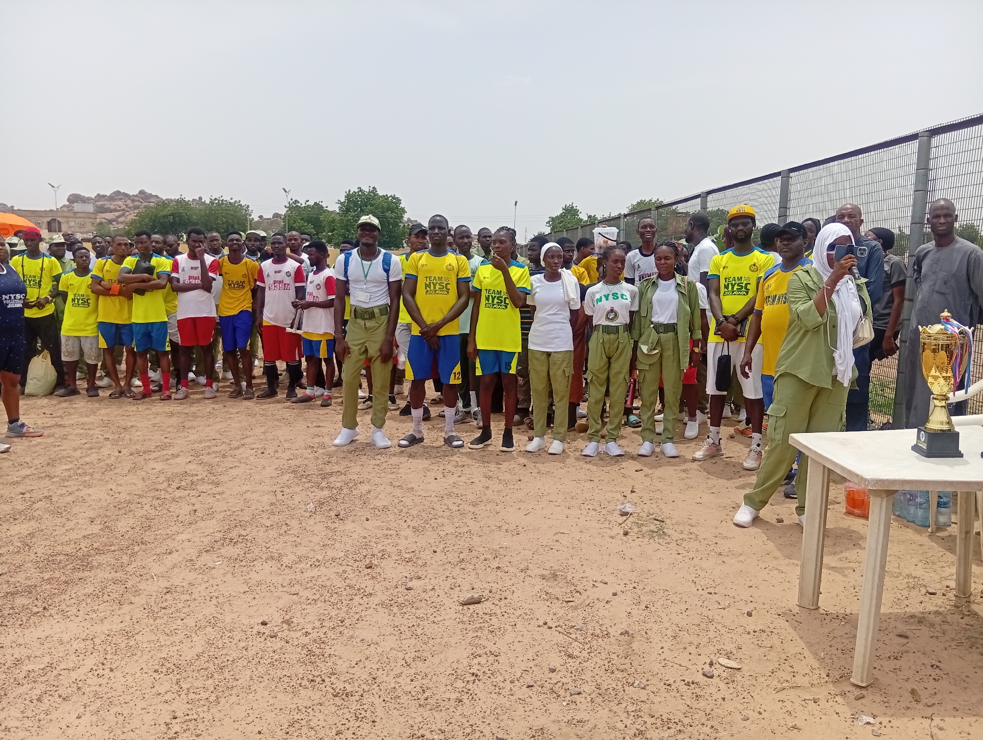 In Dutse, Nigeria, a group of individuals, likely participants in a National Youth Service Corps (NYSC) event, are gathered outdoors. The majority are dressed in sportswear, predominantly yellow t-shirts with "TEAM NYSC JIGAWA" printed on them, and various colored shorts or trousers. Some are wearing the distinct khaki uniform associated with the NYSC program.
The scene appears to be a sports event or award ceremony, suggested by the presence of a prominent trophy on a table in the foreground. The ground is a sandy, dirt surface, with scattered small stones. In the background, a chain-link fence separates the area from more distant structures and trees. Rocky hills are visible in the far distance, hinting at a semi-arid environment.
The sky is overcast, suggesting either early morning or a cloudy day. The individuals are mostly looking towards the camera, with some engaged in conversation or holding phones. Their expressions are neutral to pleasant, indicating a communal gathering. The overall atmosphere seems organized and possibly celebratory. Visible text on the shirts identifies them as "TEAM NYSC JIGAWA" and "NYSC". The trophy also has visible text, though it is not entirely legible. The presence of water bottles on the table suggests a hot climate or strenuous activity.
