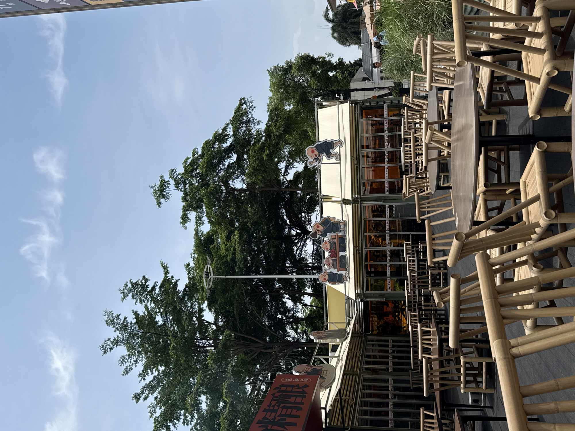 The image captures an outdoor dining area, likely associated with a restaurant or cafe, under a clear, bright blue sky with faint wispy clouds, indicating daytime. The scene suggests a prepared but currently unoccupied establishment in Haizhu District, China.
In the foreground, an array of tables and chairs crafted from light-colored bamboo are neatly arranged on a dark, paved surface. These chairs feature characteristic bamboo slats and frames, appearing robust and traditional.
Behind the seating, a modern building with large glass windows forms the main structure of the establishment. Adorning its awning or roofline are several decorative, cartoon-like cutouts depicting figures, possibly monks or traditional characters, in various poses such as sitting around a table, kneeling, or standing. A tall, thin pole with a circular ornament on top is positioned in front of a prominent, large tree with dense green foliage that dominates the left side of the frame.
A vertical red sign is partially visible in the lower left, displaying prominent Chinese characters. The most legible characters are "禅" (Chán, meaning Zen) and "鲜" (xiān, meaning fresh or delicious), suggesting a restaurant name or theme such as "Zen Fresh" or "Zen Delicacy." Smaller, indistinct text also appears on the sign.
In the background, beyond the dining area, tall green grasses and distant, blurry figures hint at a broader urban environment. The setting appears calm and ready for customers.