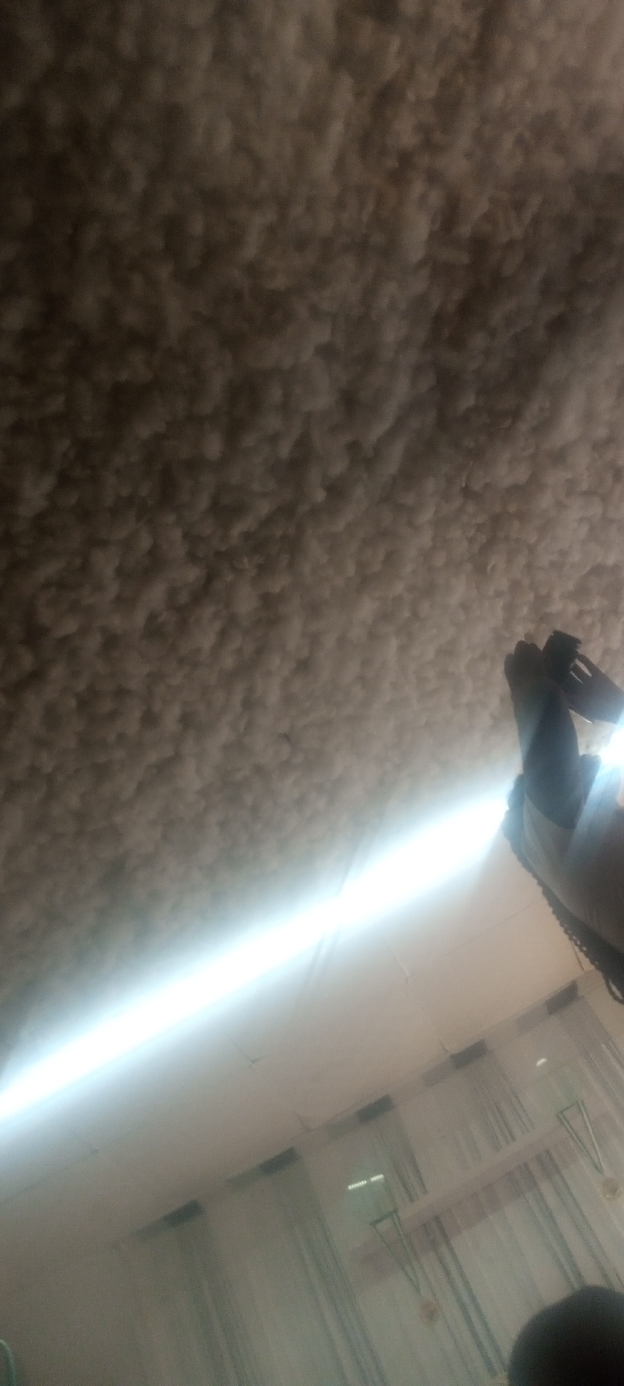 The image shows a low-angle view of a ceiling with a textured, possibly acoustic surface. A bright linear light fixture is visible in the foreground, along with some hanging objects in front of a wall. In the right side of the frame, a person is holding a mobile phone. The location is unknown, and the city is specified as "Unknown, Unknown".