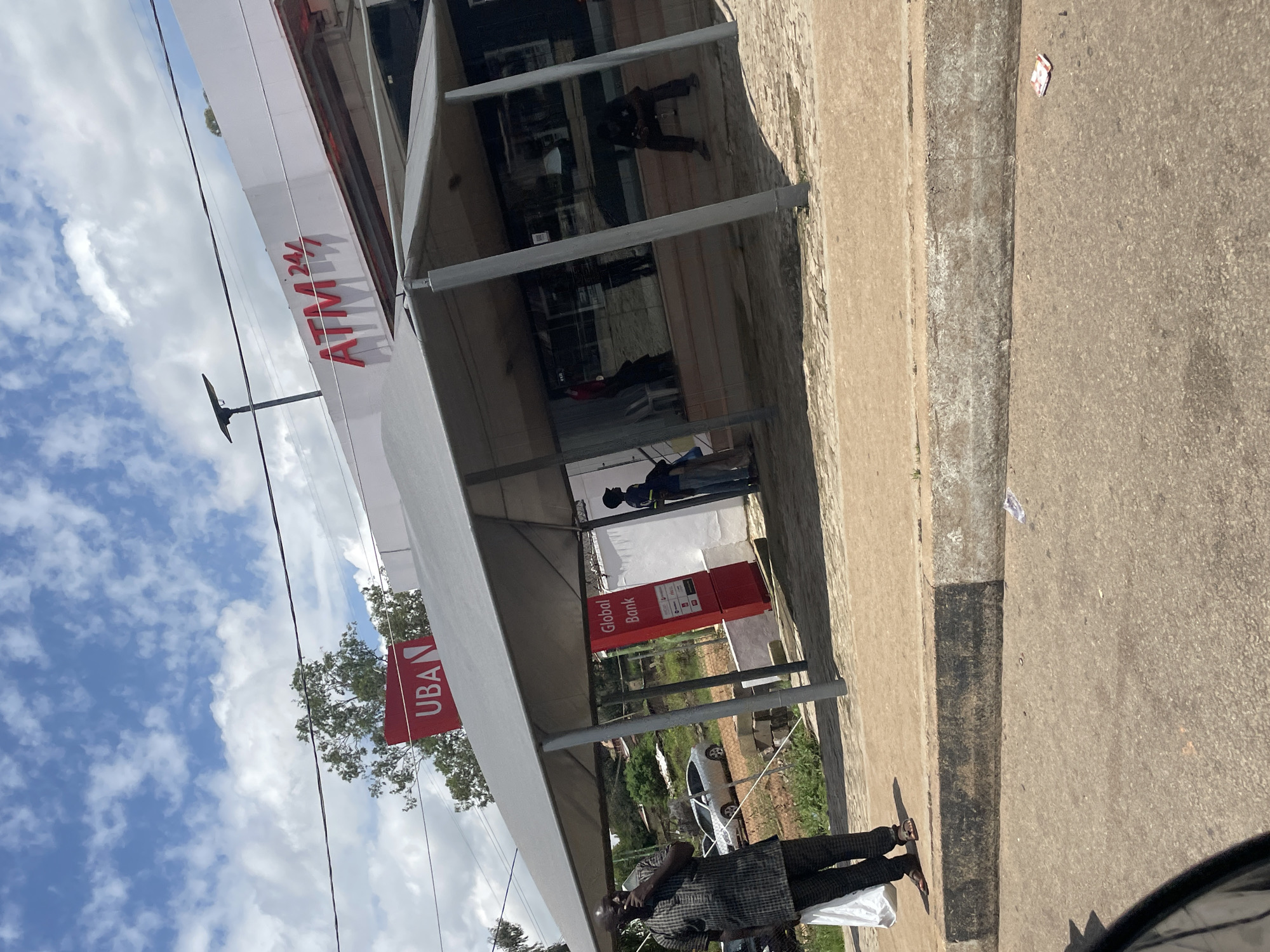 The image captures a street scene in Jos, Nigeria, featuring the facade of a United Bank for Africa (UBA) branch with an ATM. A covered outdoor seating area or shelter is set up in front of the bank. On the left, a man in traditional Nigerian attire, a patterned kaftan and trousers, stands holding a white plastic bag, his right hand near his mouth. He is wearing sandals. To the right of the man, a red sign with white lettering reads "Global Bank". Behind the shelter, reflections in the bank's windows show several people inside. Further to the right, a person in a dark shirt and light-colored pants stands near the bank entrance. The environment includes a paved sidewalk, a curb, and a road. The sky above is cloudy with patches of blue. The overall impression is of a typical day at a bank branch in a Nigerian city.