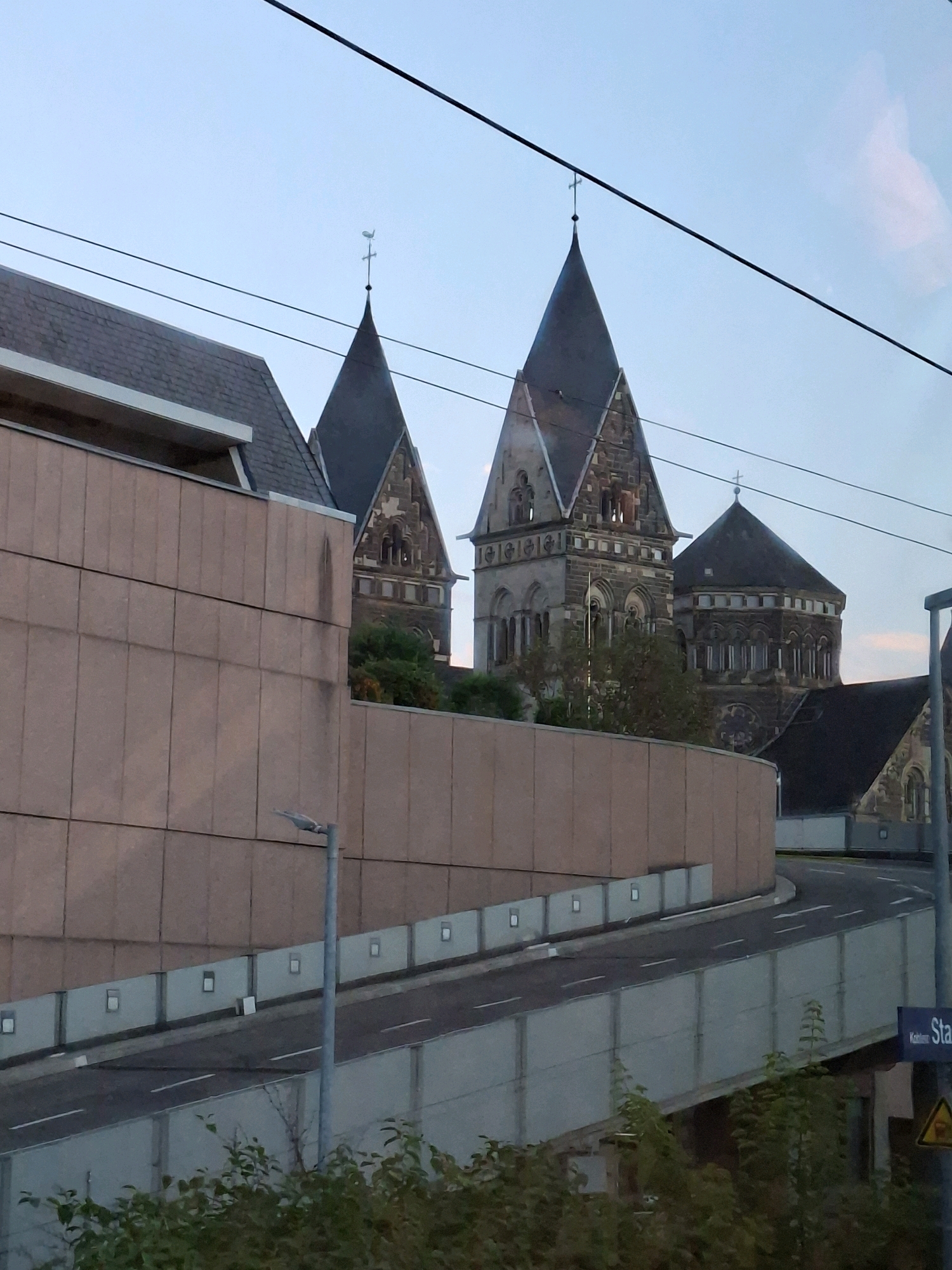 The image shows a view of the Church of St. Castor in Koblenz, Germany. The church's distinctive twin spires dominate the background. In the foreground, a modern building with a tan-colored facade is visible, partially obscuring the view of the church. A curved roadway with a light gray barrier runs in front of the modern building, separating it from a patch of greenery at the bottom of the frame. The sky is overcast, and the lighting suggests it is either early morning or late evening. A street sign with the word "Sta" visible is positioned on the right side of the image.