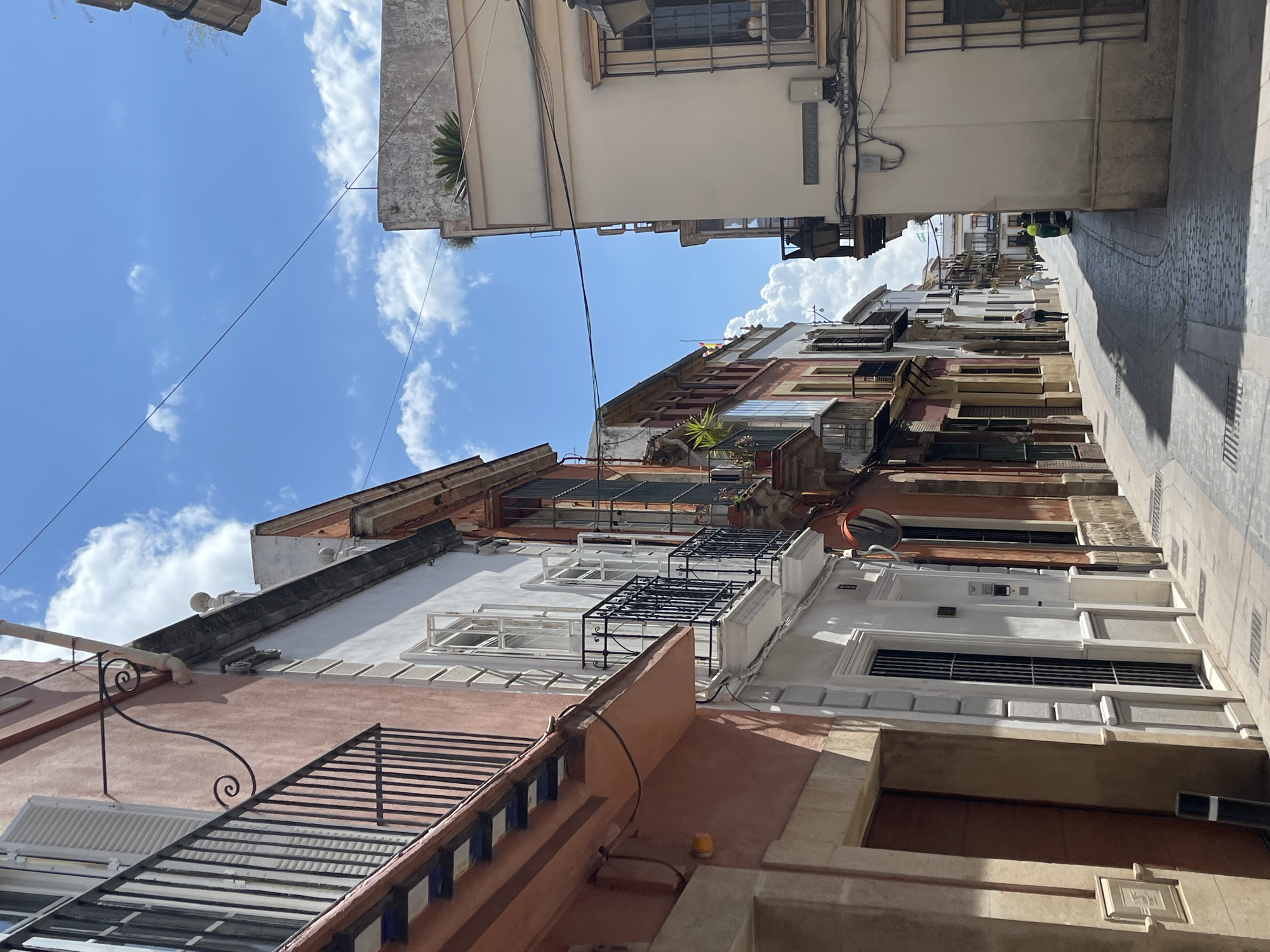 An alleyway in Jerez De La Frontera, Spain, is viewed from a low angle, featuring buildings with a mix of architectural styles in shades of white, brown, and orange. The buildings have various balconies, windows, and decorative elements. The sky is blue with scattered clouds. A pedestrian is walking down the alleyway. Overhead wires are strung between the buildings.
