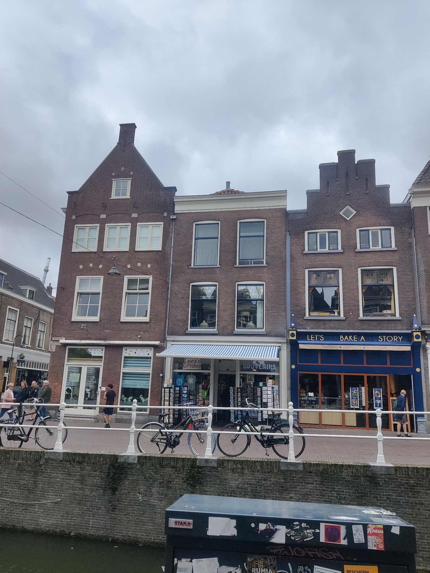 The image shows a street scene in Delft, Netherlands, with traditional Dutch brick buildings lining a canal. The sky is overcast. In the foreground, a brick wall separates the canal from the sidewalk. Several bicycles are parked along a white railing on the sidewalk.
On the right side of the street, a bakery with a blue facade displays the text "LET'S BAKE A STORY" in gold lettering. The bakery also has signs indicating "patisserie," "viennoiserie," and "boulangerie." Next to it, a shop labeled "SOUVENIRS" has a blue and white striped awning. Further to the left, a building has "BEAUTY LOUNGE" written on its window. Several people are walking on the sidewalk.
The architecture of the buildings is characteristic of Delft, with gabled roofs and multi-pane windows. The building on the far left has a sign that reads "WYNHAVEN". The overall impression is a lively street scene in a historic Dutch city.