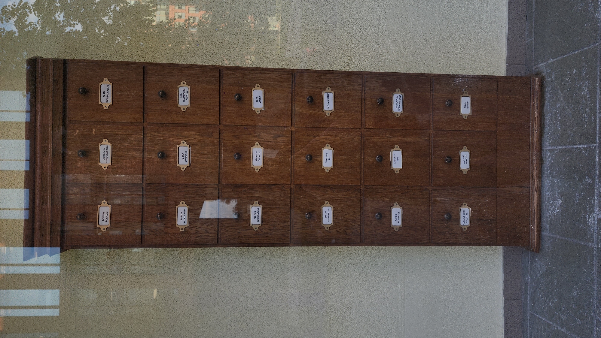 The image shows a tall, wooden cabinet with multiple drawers, each labeled with a brass plaque. The cabinet appears to be an antique apothecary cabinet. The labels on the drawers are in Latin and indicate various substances, such as "ACIDUM BORICUM," "ZINCOXYDUM HYDROCHLORIDUM," and "CARBO ACTIVATUS." The cabinet is placed against a textured yellow wall, and the floor is paved with grey stone tiles. Reflections of a window with vertical blinds and a tree with foliage are visible on the glass front of the cabinet. The scene is lit by natural light, casting soft shadows. The overall impression is one of an old-fashioned storage unit, possibly in a museum or a historical pharmacy.
The provided context indicates that the location is in Moscow, Russia, but this information is not visually evident from the image itself. The image does not contain any people, actions, or interactions. The environment is static and focuses solely on the cabinet and its immediate surroundings.