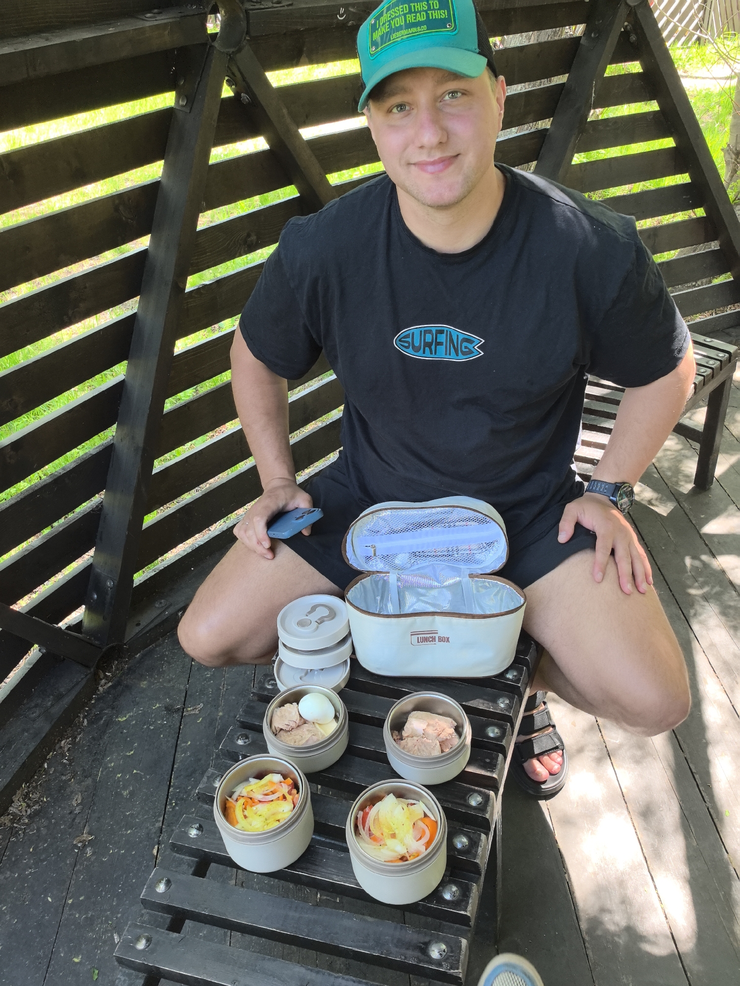 A young man wearing a black t-shirt with a "SURFING" logo and a green baseball cap that reads "I DRESSED THIS TO MAKE YOU READ THIS!" kneels on a wooden bench. He is holding a blue smartphone in his right hand. In front of him are several round, stacked food containers and an open lunchbox. The lunchbox is light beige with a white interior and a zipper. The food containers hold what appears to be boiled eggs, canned meat, and chopped vegetables. The man is in an outdoor setting, within a structure made of dark wood slats, with green grass visible through the gaps. This scene is located in Yelizovo, Russia.