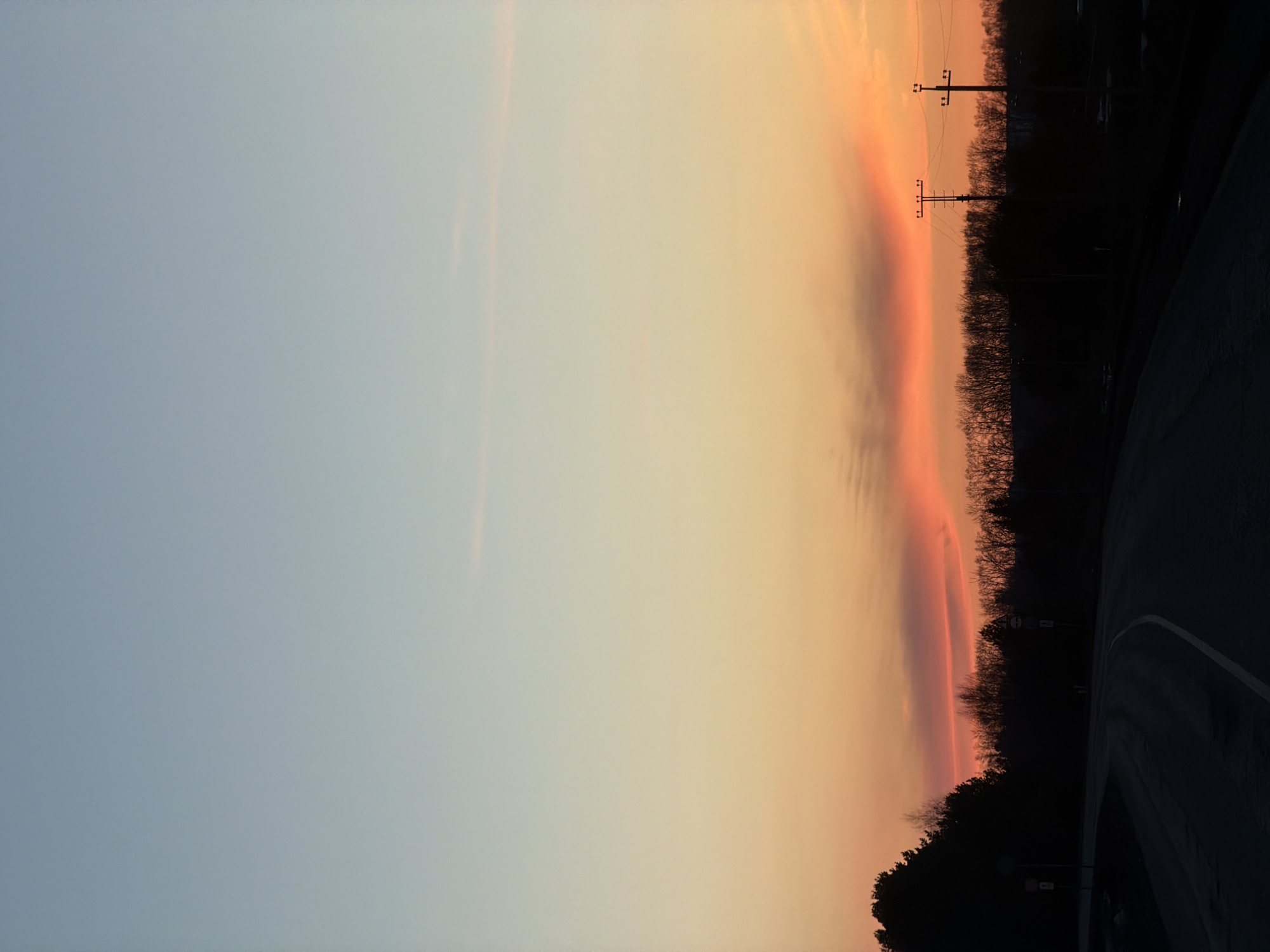 The image shows a sunset over a road near Gory, Russia. The sky transitions from light blue to orange, with a distinct pinkish band near the horizon. Silhouetted trees and power lines are visible on the right. The road curves into the foreground, disappearing into shadow.