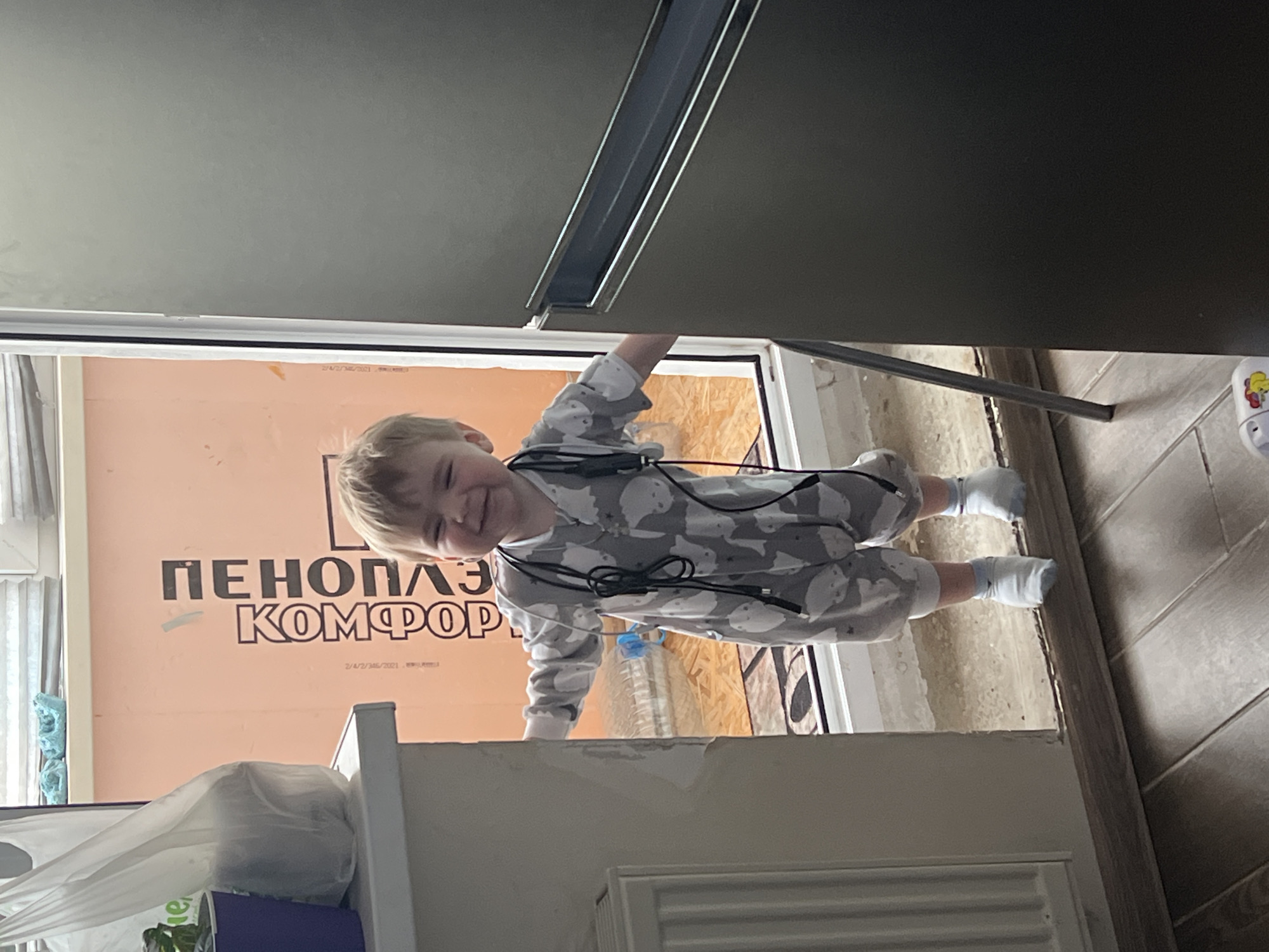 A young child is peering into the camera from between a refrigerator and a counter. The child is wearing a gray and white pajama set and white socks. Cords are draped around the child's neck. Behind the child is a window and an exterior wall insulated with rigid foam insulation, printed with the word "ПЕНОПЛАСТ" and the word "КОМФОРТ" in large, black letters. The foam insulation also has a date printed on it: "2/4/2021". The floor is tiled. The scene is inside a building, likely in Noviy, Russia, based on the Cyrillic script.