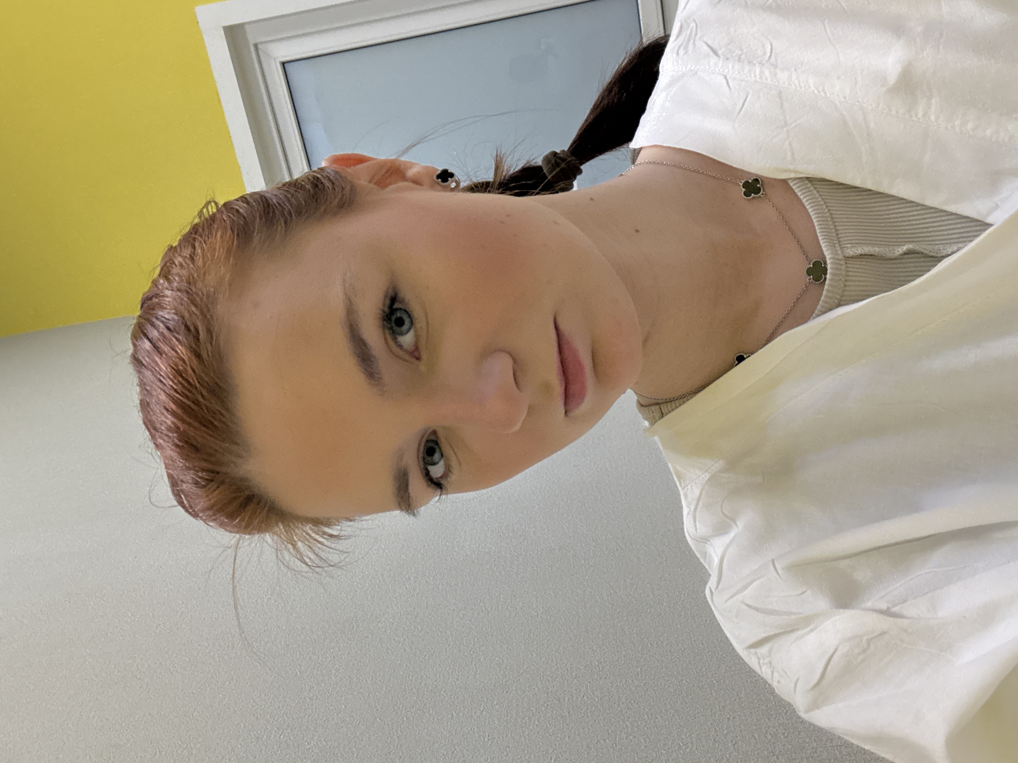 The image is a close-up, low-angle shot of a woman with light skin, blue eyes, and reddish-brown hair pulled back into a braid. She is wearing a white, collared shirt, revealing a light-colored, possibly ribbed undershirt and a silver necklace with clover-shaped pendants. She also has a clover earring. The background includes a window with a white frame and what appears to be a yellow wall in the upper left. The lower left background is a light gray wall. There is no location in the city of Unknown, Russia visible in the image.