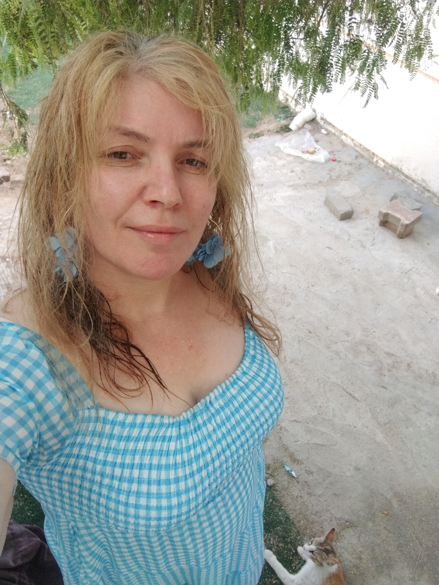 A woman with blonde, slightly wet hair, wearing a blue and white checkered dress and blue flower earrings, is taking a selfie outdoors. She's likely on a patio or paved area, evidenced by concrete blocks and pavement. A cat is visible in the lower right corner. The location is Al Mas'oudi, United Arab Emirates, although specific evidence within the image to confirm this location is limited. Green foliage of a tree or bush is visible in the background. The environment is bright with natural light. A plastic bag and other debris are on the pavement.