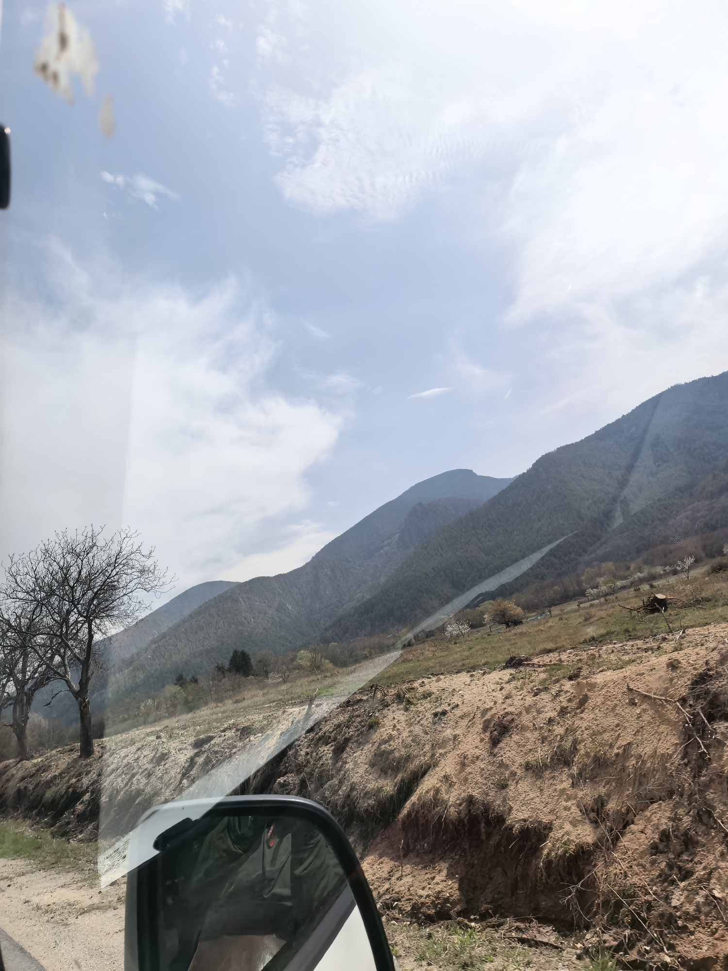The image shows a landscape seen from inside a vehicle. In the foreground, the side mirror and the edge of the car's window frame are visible. The immediate ground outside the car appears to be a dirt road or path. Beyond that, there is a steep dirt embankment. Trees with bare branches stand near the road. In the background, rolling hills and mountains extend toward the horizon under a partly cloudy sky. The location is in the city of Unknown, Unknown.