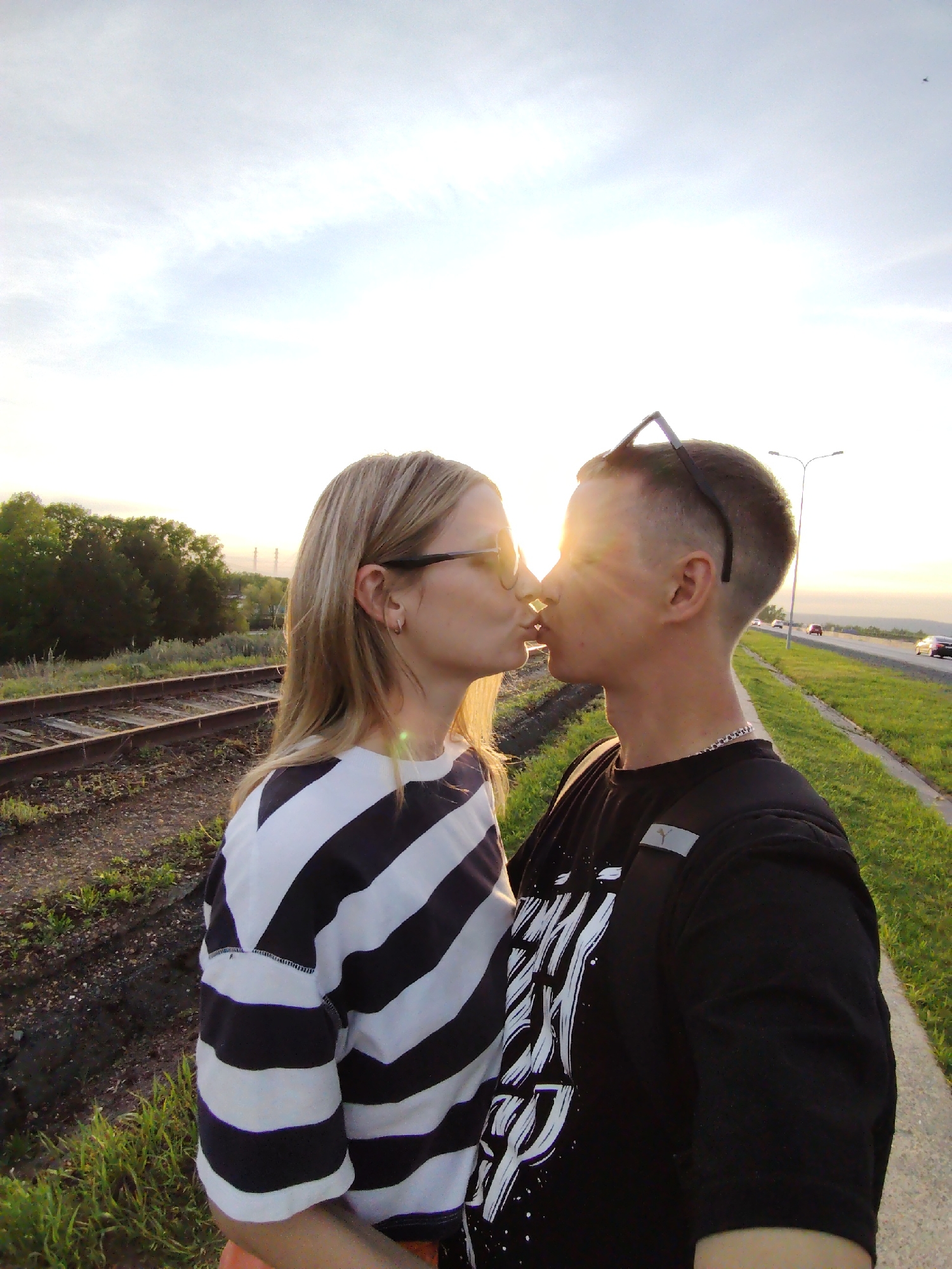 A man and woman are kissing outdoors. The woman has blond hair, wears glasses, and a black and white striped shirt. The man wears a black t-shirt with white graphics. Sunglasses are perched on his head. They are standing next to a sidewalk and a grassy area adjacent to a highway with passing cars. Parallel to the sidewalk are railroad tracks. Trees are in the background. The sun is low in the sky, creating a bright glow. The location is Chaikovsky, Russia.