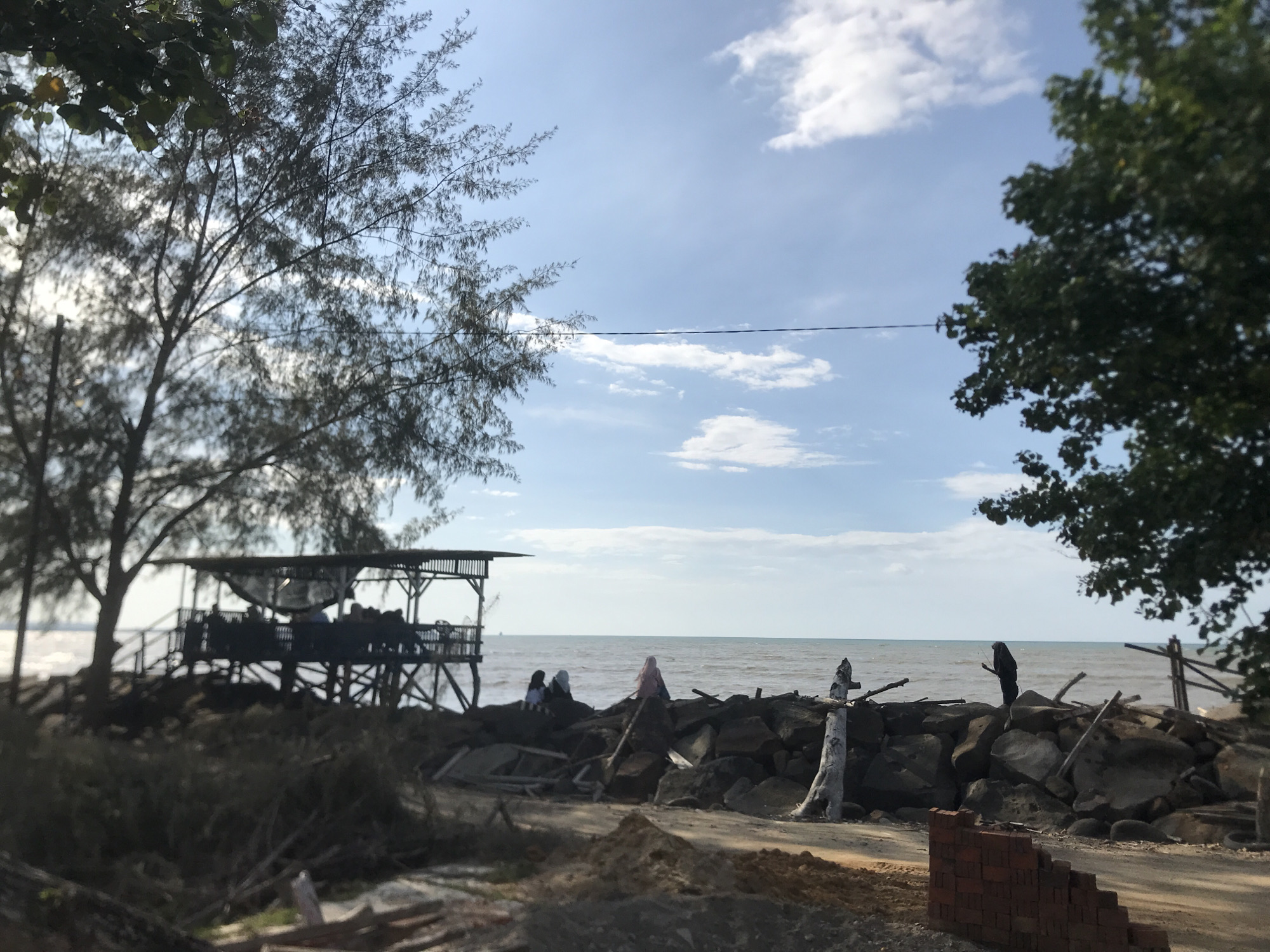 The image depicts a coastal scene in Lancok, Indonesia, with a muted blue sky and calm sea. A wooden structure, resembling a pier or observation platform, is built over the water to the left. Several people are on this structure. Further to the right, individuals, one wearing a pink hijab, are gathered on a rocky embankment. A solitary figure stands on the rocks to the right. In the foreground, piles of dirt, debris, and stacked bricks are visible. Trees frame the scene on both sides. An electrical wire crosses the sky.