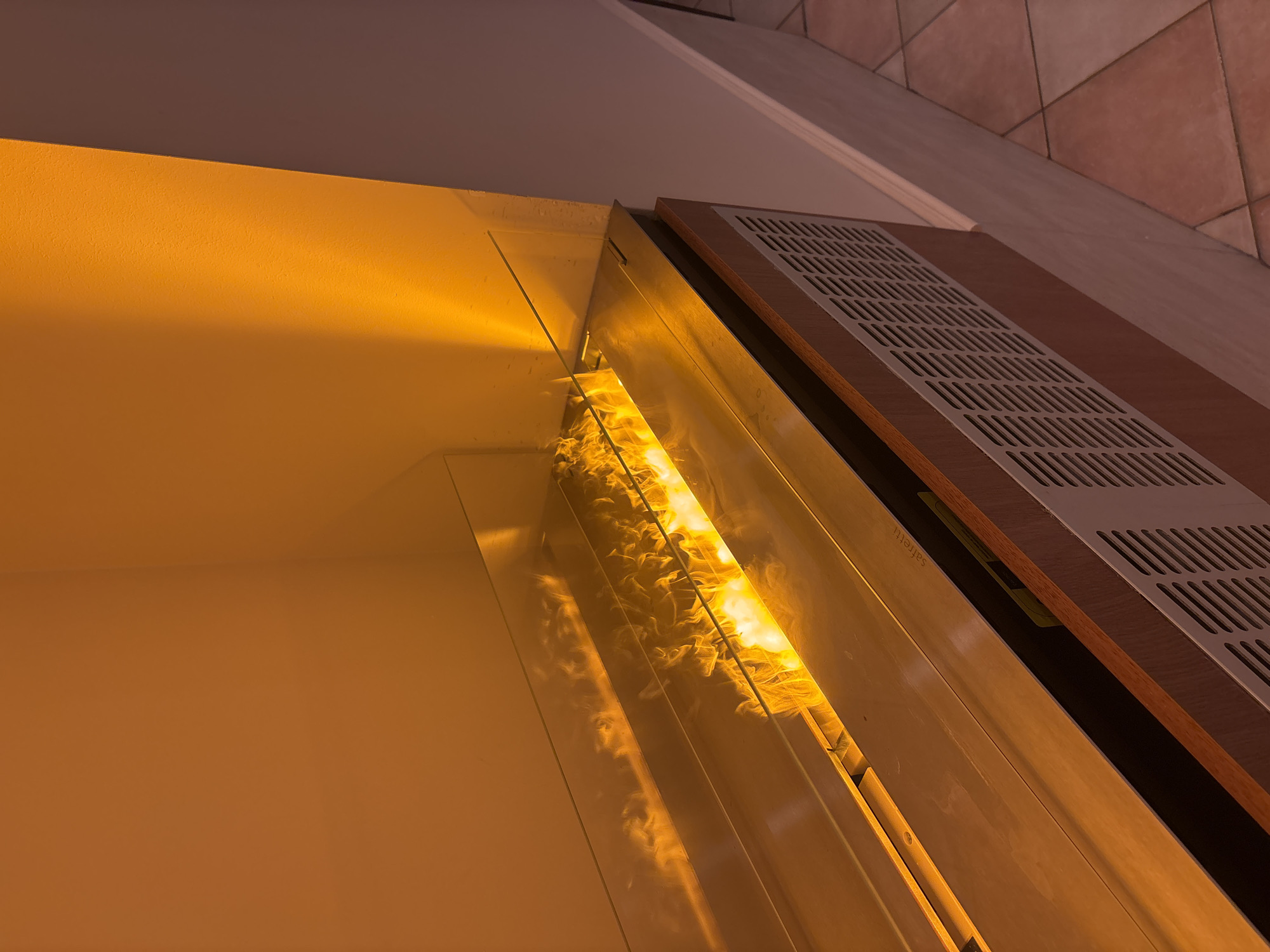 The image shows a close-up, interior shot of a modern bioethanol fireplace. The fireplace is integrated into a wall or built-in unit, with a polished metal surround and a glass wind guard. The flames are a bright orange and yellow, casting a warm glow on the surrounding surfaces. The wall behind the fireplace is painted a warm, orangey-yellow, with a distinct shadow line indicating the corner of the room. To the right of the fireplace, a neutral-colored wall curves away from the viewer, meeting a tiled floor in a reddish-brown hue. The lower portion of the fireplace unit features a wooden trim and a white metal grate, likely for ventilation. The scene is well-lit, with the primary light source appearing to be the fire itself and possibly ambient lighting. There are no people or animals visible in the image. The location is specified as Razliv, Bulgaria.