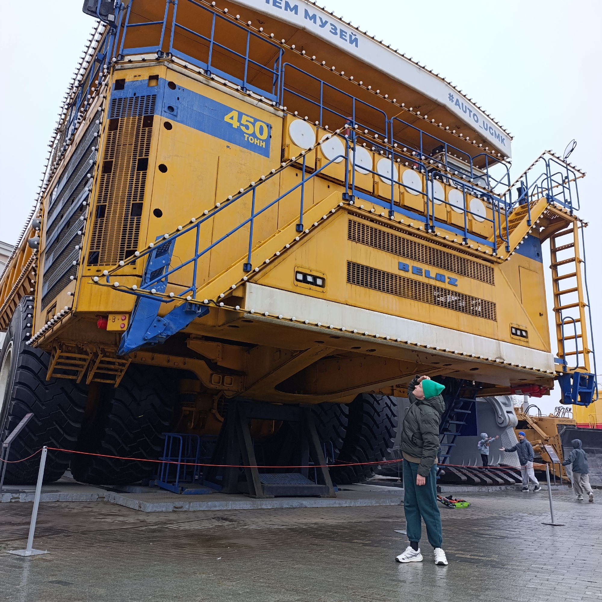 An immense, yellow and blue BelAZ mining dump truck dominates the scene at an outdoor exhibition in Verkhnyaya Pyshma, Russia. The vehicle, an impressive example of heavy machinery, is adorned with numerous small white string lights, giving it a striking appearance. Blue railings and steps provide access points up its colossal body.
Visible text on the truck includes "450 ТОНН" (450 TONN), highlighting its massive carrying capacity, and "BELAZ," identifying the manufacturer. Further along the top edge, partial text reads "ЕМ МУЗЕЙ" (suggesting "MUSEUM") and "#AUTO_UGMK," indicating its likely display at an automotive museum associated with the Ural Mining and Metallurgical Company.
In the foreground, a young boy in a green beanie and dark jacket looks up in awe at the truck's towering scale. Other visitors, including two adults and a child, are visible further back near the rear of the vehicle, also observing the exhibit. The ground is wet, suggesting recent rain or drizzle, and the sky is overcast, providing soft, diffused lighting. A red rope barrier marks off a section of the display. The truck rests on large support structures, emphasizing its static exhibition role. A small sign in the background bears the text "ПРОЕКТОР" (PROJECTOR). The overall impression is one of wonder at the sheer size and engineering of the industrial vehicle.