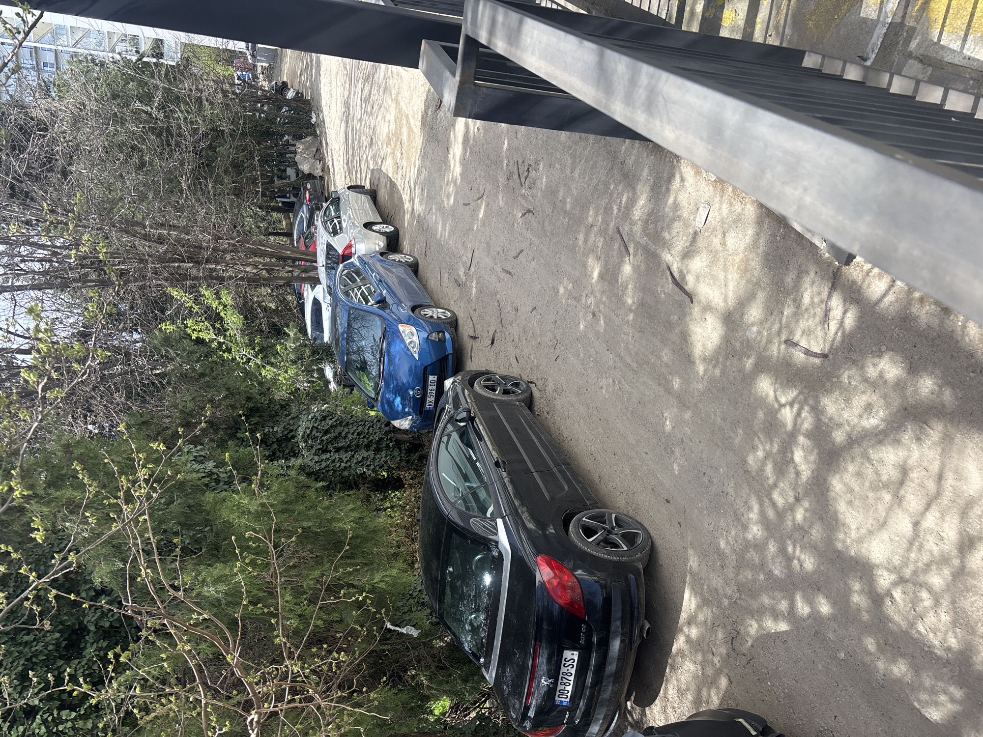 A street in Tbilisi, Georgia, shows a row of parked cars under trees. A black Peugeot 207 CC is parked closest to the camera, with a visible license plate "GG-878-SS." Further along, a blue car with a license plate "KK-596-DD" is parked. Several other vehicles are parked behind it, partially obscured by the trees. The ground is unpaved. A metal railing or bridge is visible above. Sunlight creates shadows across the street.