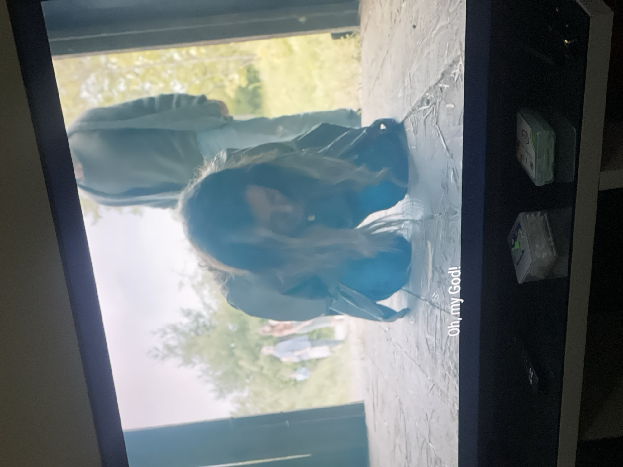 The image shows a television screen displaying a scene with a person kneeling, facing down with their head close to the ground. The person is wearing a jacket and has dark hair. In the background, figures are visible standing outdoors in a green, tree-filled environment. Text on the screen reads "Oh, my God!". The setting appears to be outside an open doorway or structure. The TV is on a shelf with a remote control and various objects. The image provides no information indicating that this scene took place in Abuja, Nigeria, or any specific location.