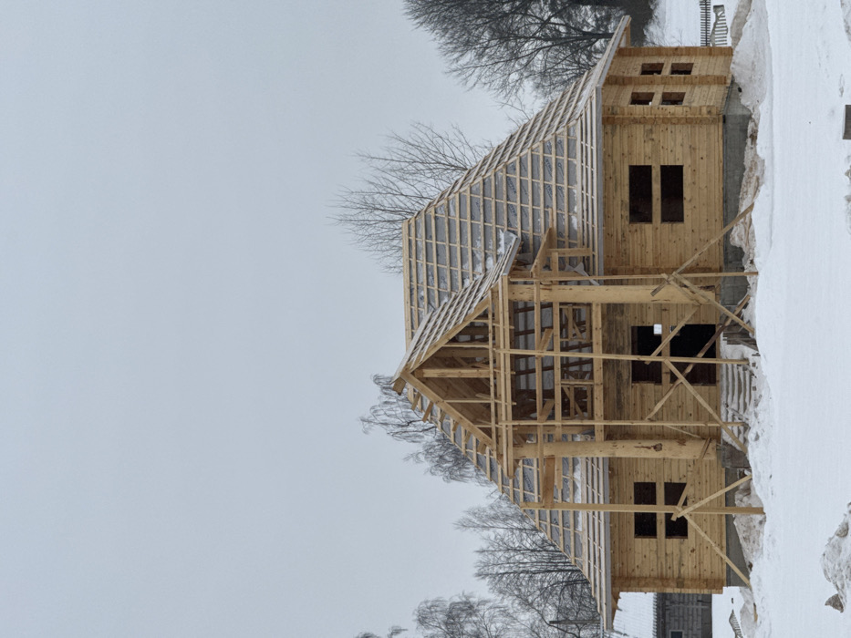 The image depicts a two-story wooden house under construction in Крэўскі сельскі Савет, Belarus, under an overcast sky during winter. The ground is covered in white snow. The house is constructed from light-colored logs, with an exposed gray concrete foundation. Multiple rectangular openings serve as provisional windows and a main entrance, lacking frames or glass. A complex, multi-gabled roof structure is evident, featuring exposed wooden rafters and a partial protective underlayment, lightly dusted with snow. A prominent A-frame gable with visible wooden framing stands over the front entrance area. Temporary wooden support beams are positioned around the front porch structure. In the background, bare deciduous trees are visible on both sides of the house, and a small, low-lying snow-covered wooden structure is partially visible to the far left. A snow-covered fence line and a gentle upward slope of snow are visible to the right. No individuals are present.