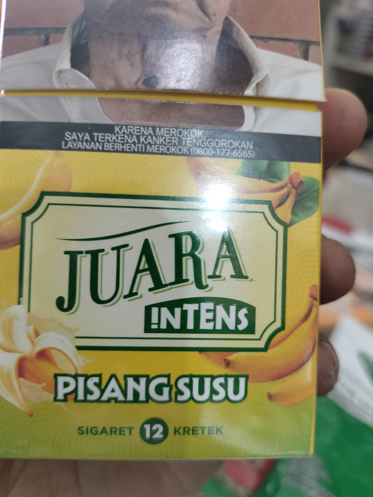 The image shows a close-up of a cigarette pack featuring a graphic of bananas and text in Indonesian. The pack is yellow and green with a black band at the top. The text on the black band reads "KARENA MEROKOK SAYA TERKENA KANKER TENGGOROKAN LAYANAN BERHENTI MEROKOK (0800-177-6565)", which translates to "BECAUSE I SMOKE I HAVE THROAT CANCER QUIT SMOKING SERVICE (0800-177-6565)". The brand name "JUARA" is prominently displayed in large green letters, with "INTENS" written below it. Below that, "PISANG SUSU" is written, meaning "Banana Milk". At the bottom, "SIGARET 12 KRETEK" indicates it is a clove cigarette with a strength of 12. The image is captured at an angle, and a person's hand and thumb are visible on the right side, partially obscuring the cigarette pack. The background is blurred, suggesting an indoor setting, possibly a shop or a market stall, consistent with the commercial nature of the product. The specific location within the city of Meudangara, Indonesia, cannot be determined from the visual information provided in this image.