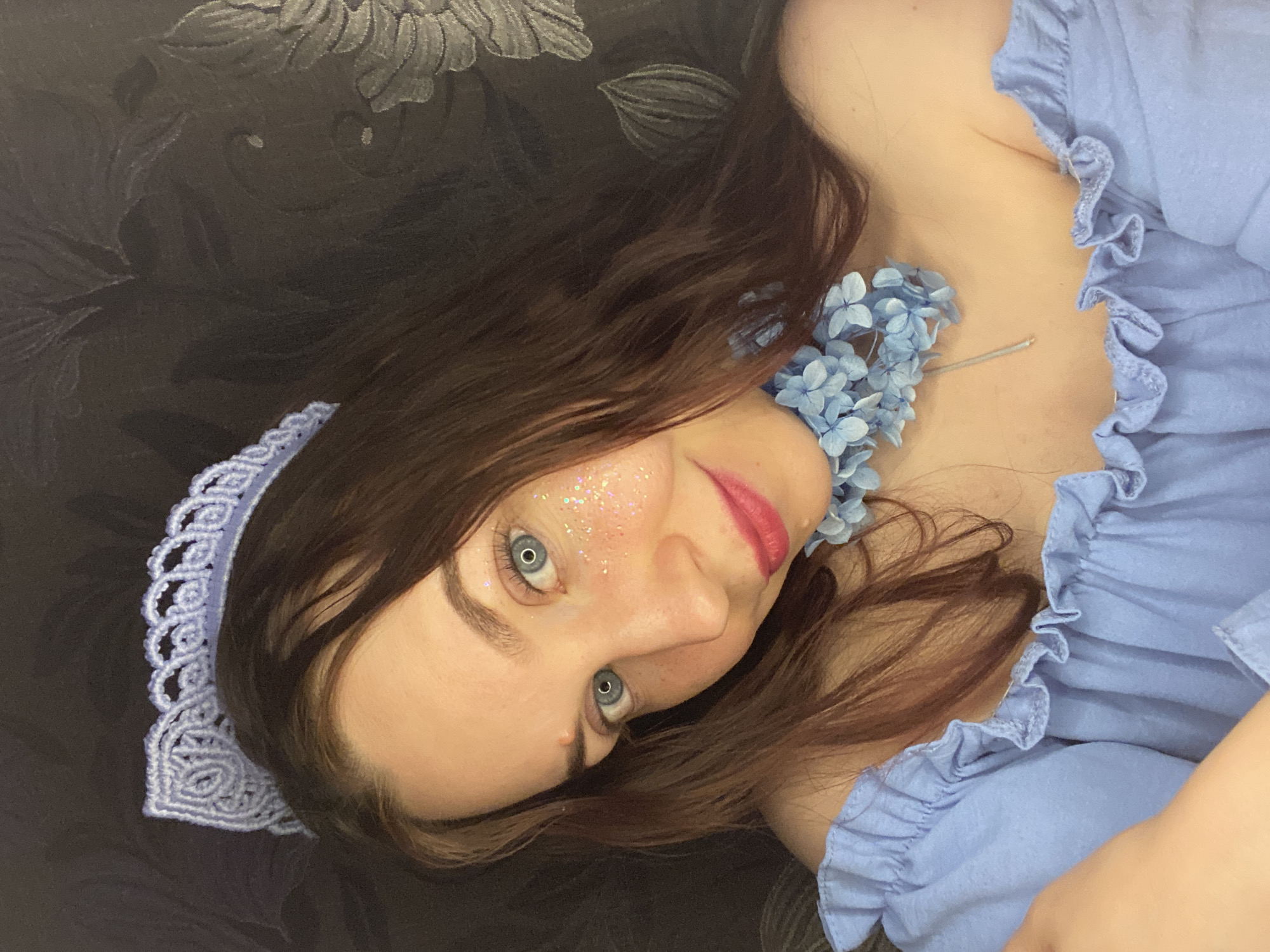 A young woman with long brown hair, fair skin, and blue eyes is lying on a dark, patterned surface. She is wearing a light blue dress with ruffled edges and a matching lace headband. Her face is decorated with glitter. A necklace of light blue flowers adorns her neck. The location of the scene in the city of Chaikovsky, Russia, is not discernible from the provided image.