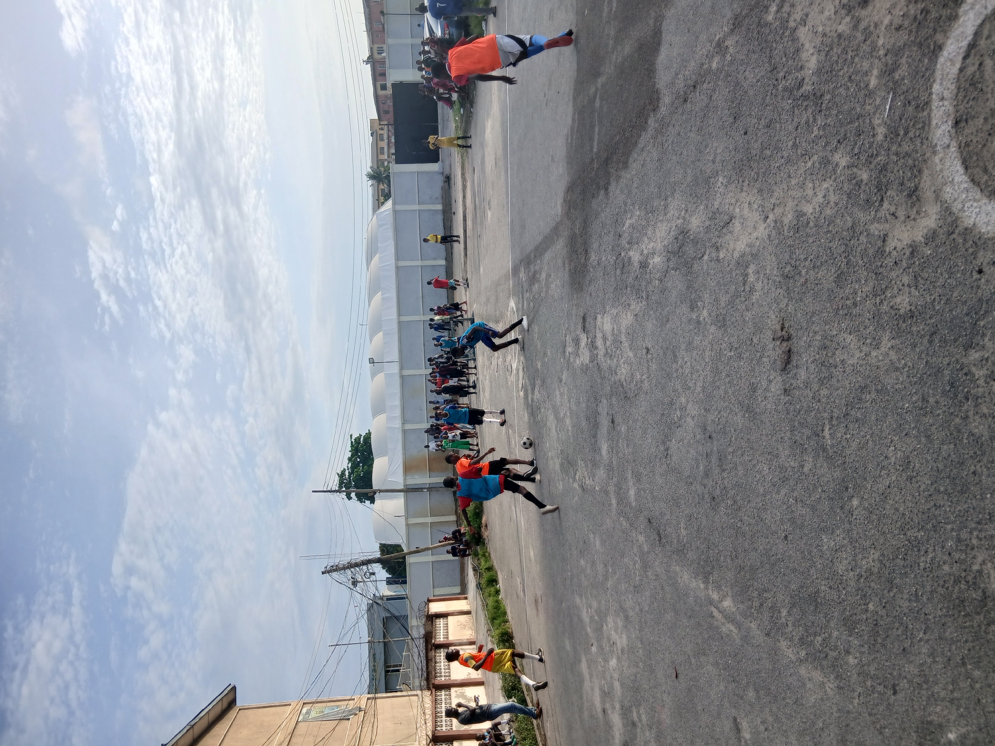 In Lagos, Nigeria, a daytime scene shows a group of people engaged in a soccer game on an asphalt court. Some individuals are wearing orange and blue jerseys. A white-walled building runs along one side of the court, and utility poles are visible near a low wall. The sky is overcast with scattered clouds. Buildings border the court, indicating an urban setting.