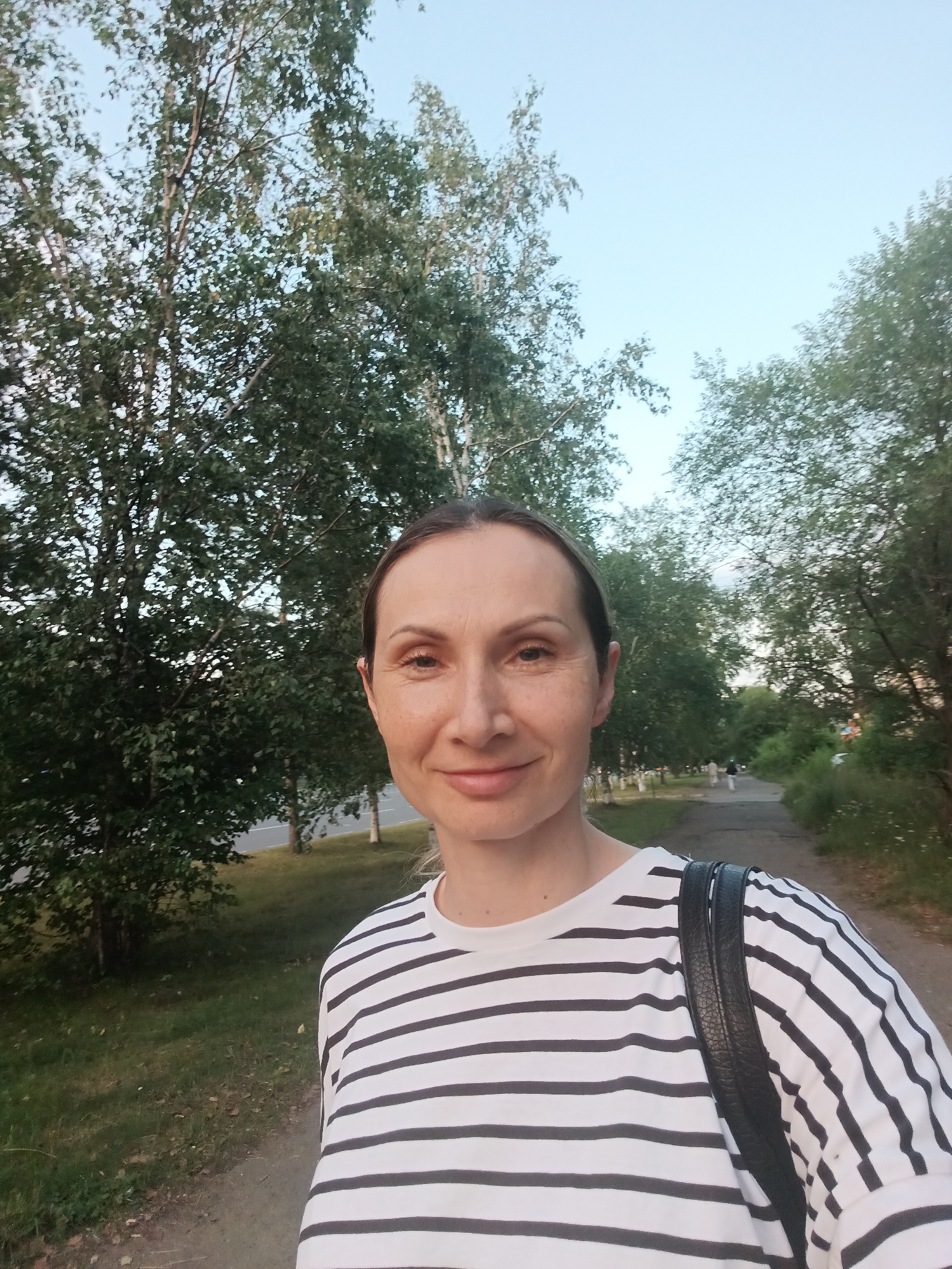 A woman is taking a selfie outdoors on a path lined with trees. She has light skin and brown hair pulled back. She is wearing a black and white striped t-shirt and a black backpack. The background shows a grassy area, a road with sparse traffic, and more trees. The sky is clear and blue. The location is on a pathway adjacent to a road in Komsomolsk-on-Amur, Russia, possibly in a park or along a residential street.